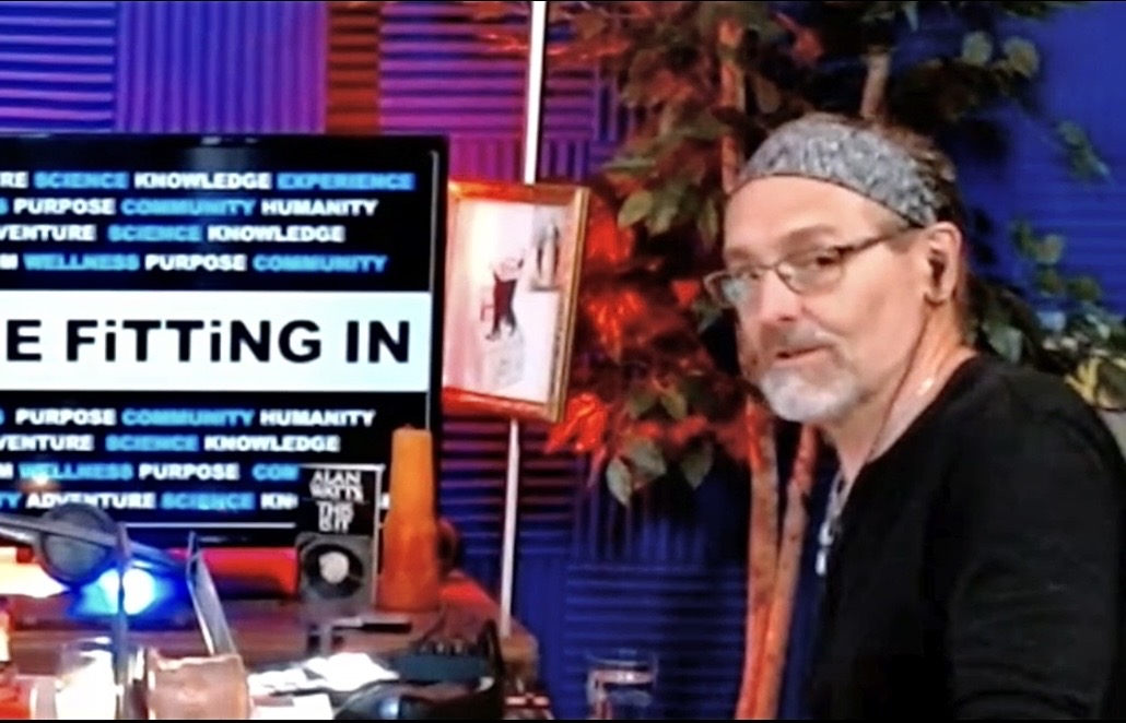 Man with glasses and headband sitting at a desk with a colorful background and a screen displaying text.