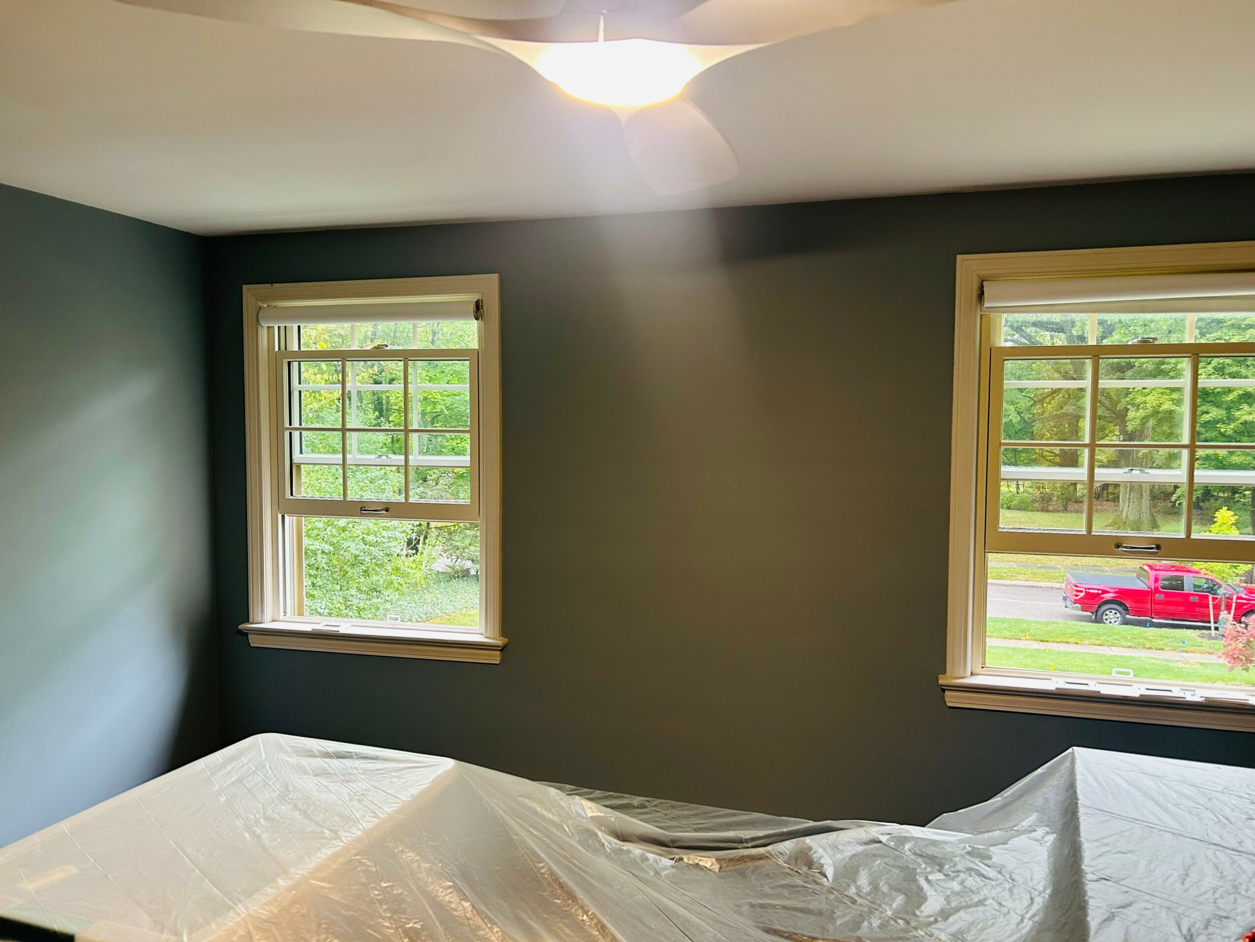 Room with two windows showing outdoor greenery and a red vehicle, ceiling light, and a covered furniture piece.