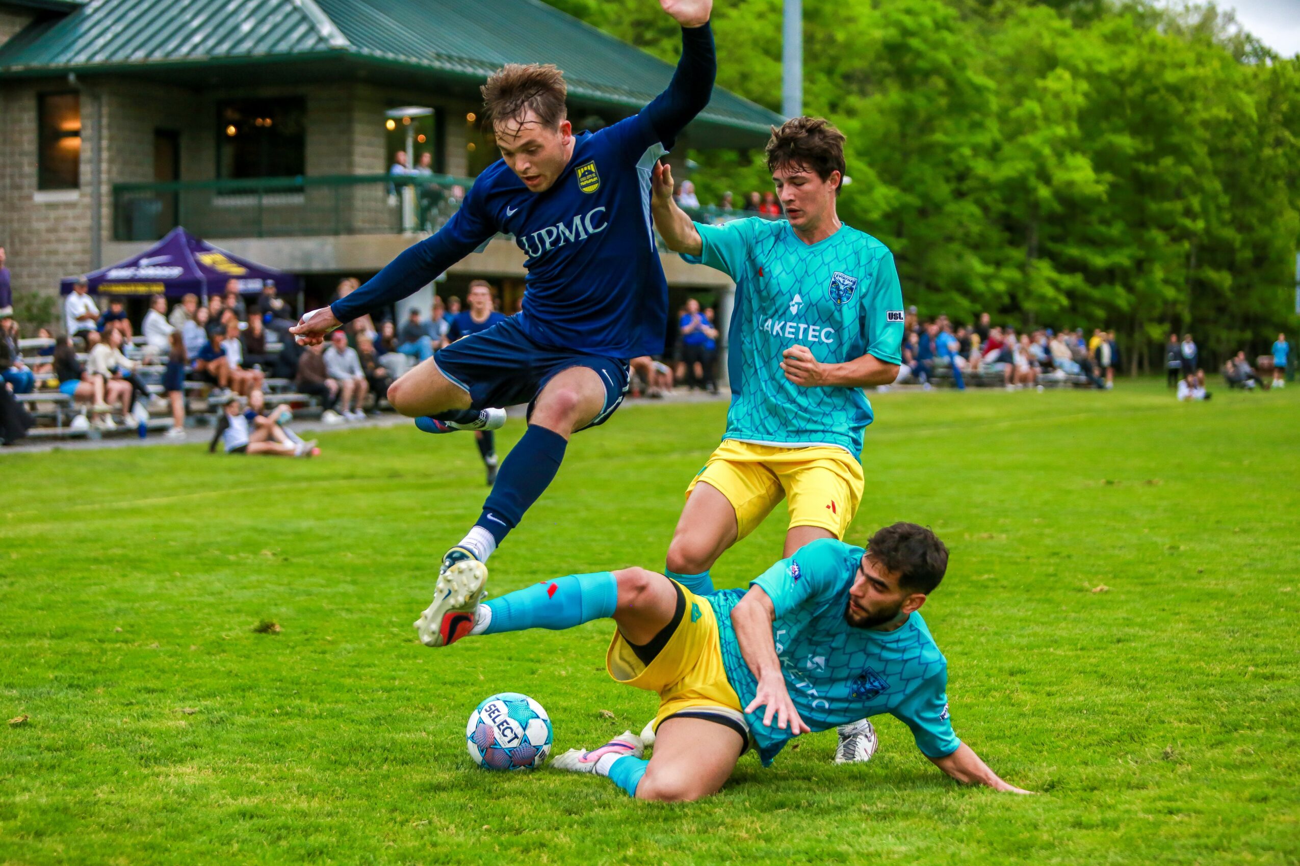 Two soccer players in blue and yellow uniforms compete for the ball on a grassy field, with spectators and trees in the background.