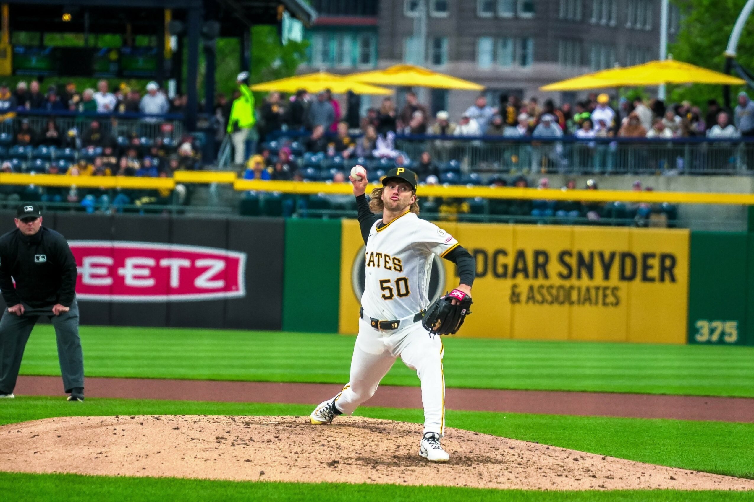 Baseball pitcher in uniform throws a pitch on the field with spectators in the background.