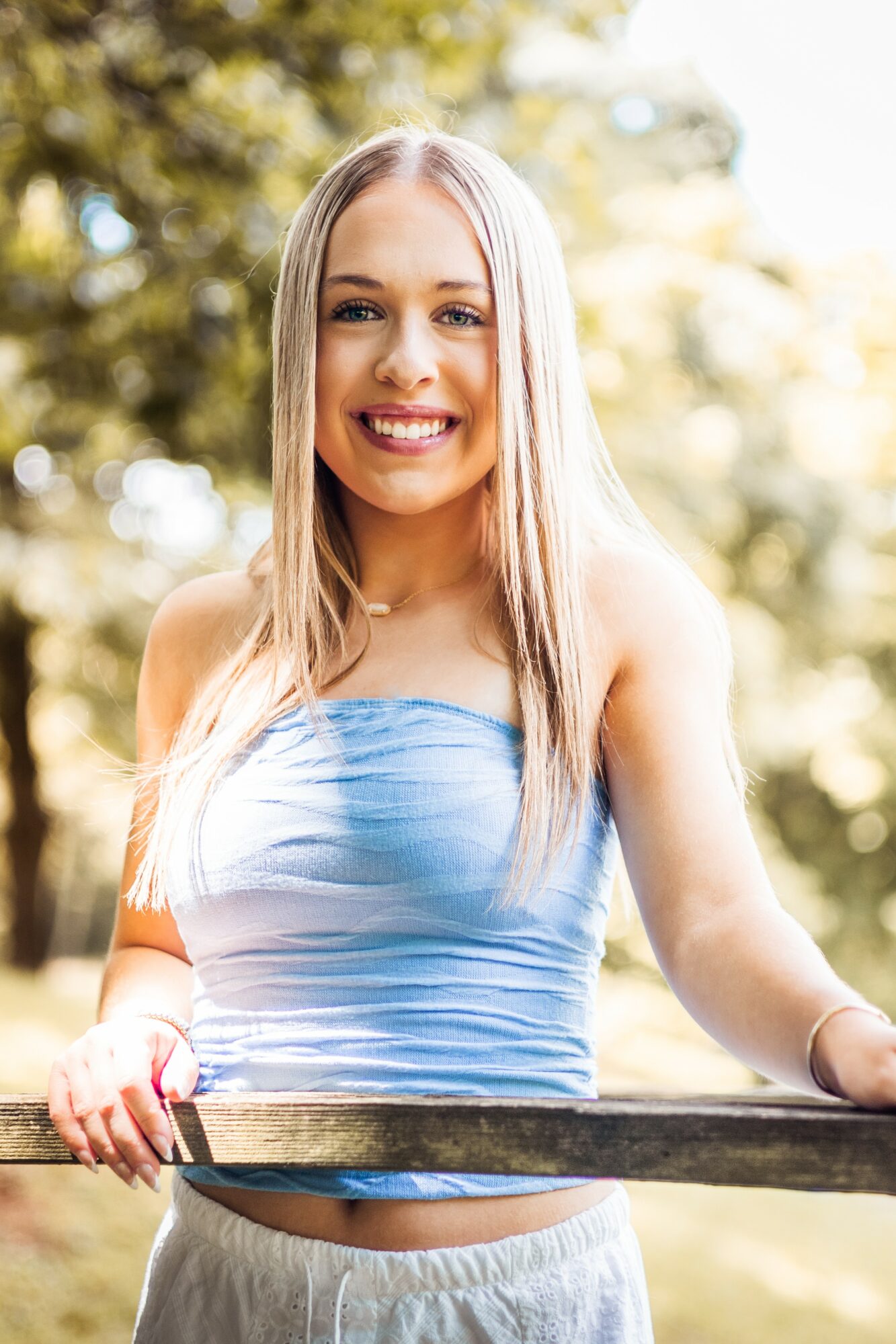 Smiling young woman with long blonde hair outdoors, wearing a blue strapless top and light-colored pants.