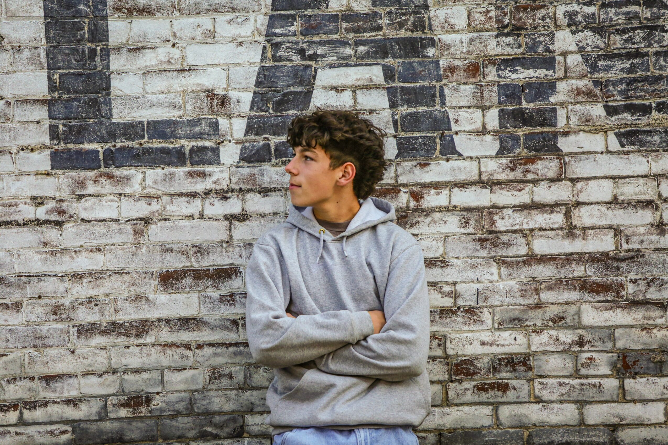 Young person with curly hair in a gray hoodie standing against a brick wall, arms crossed, looking to the side.