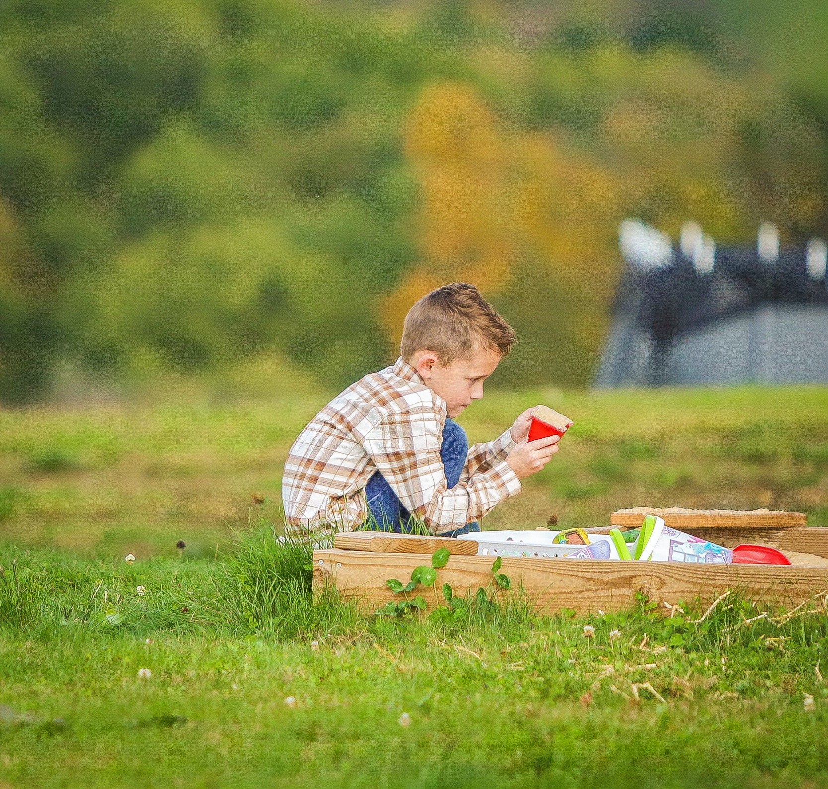 Child sitting outdoors, playing with a red toy, surrounded by grass and trees, with a blurred background of a vehicle and trees.