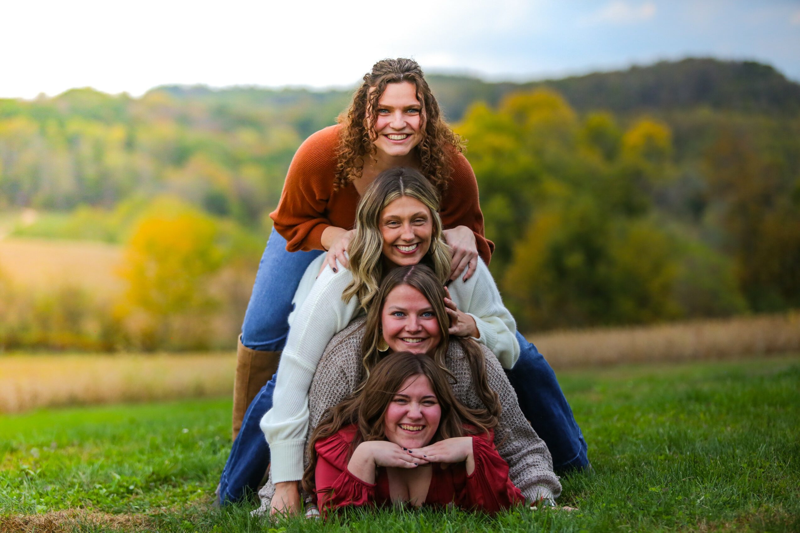 Four women stacked in a pyramid outdoors, smiling, with a landscape of trees and hills in the background.