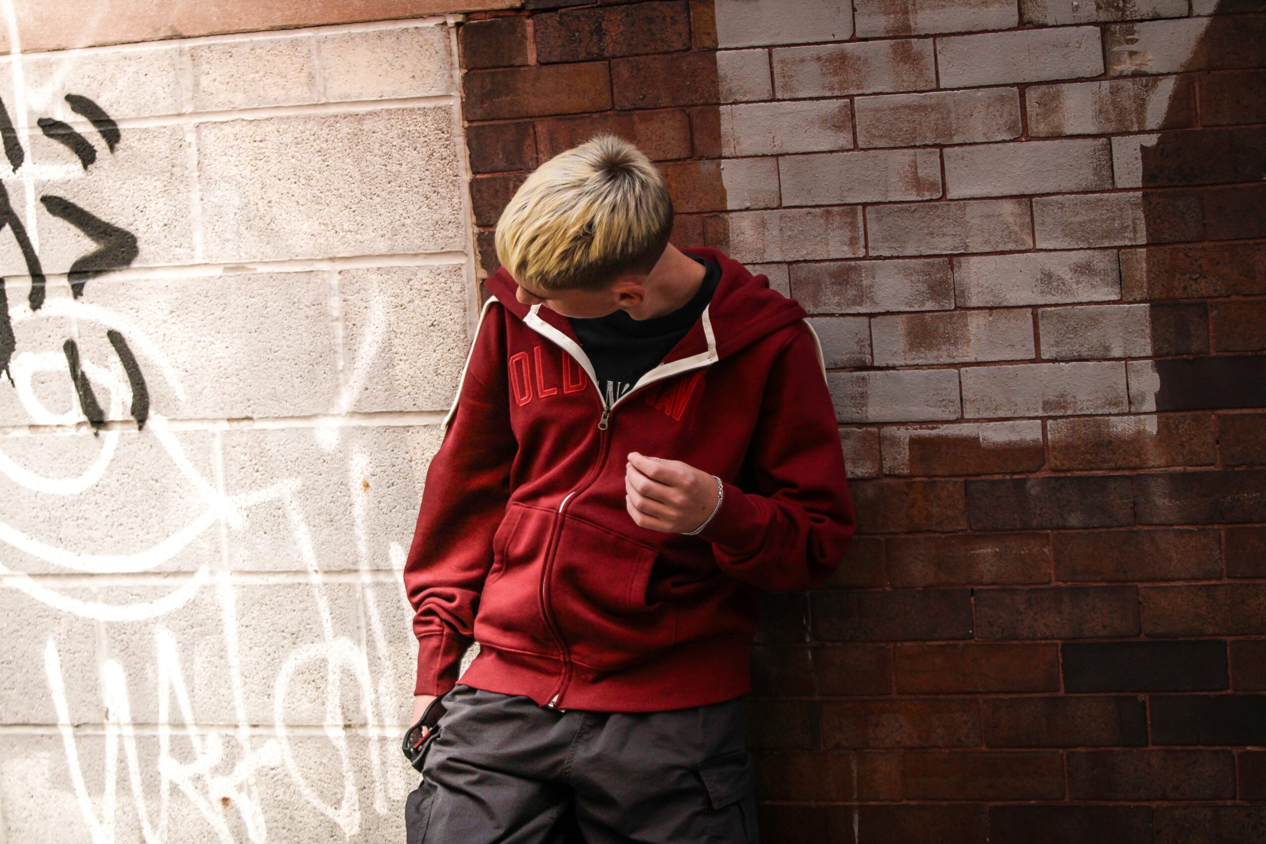 Person with blonde hair wearing a red jacket and gray pants, standing against a brick wall, looking down.