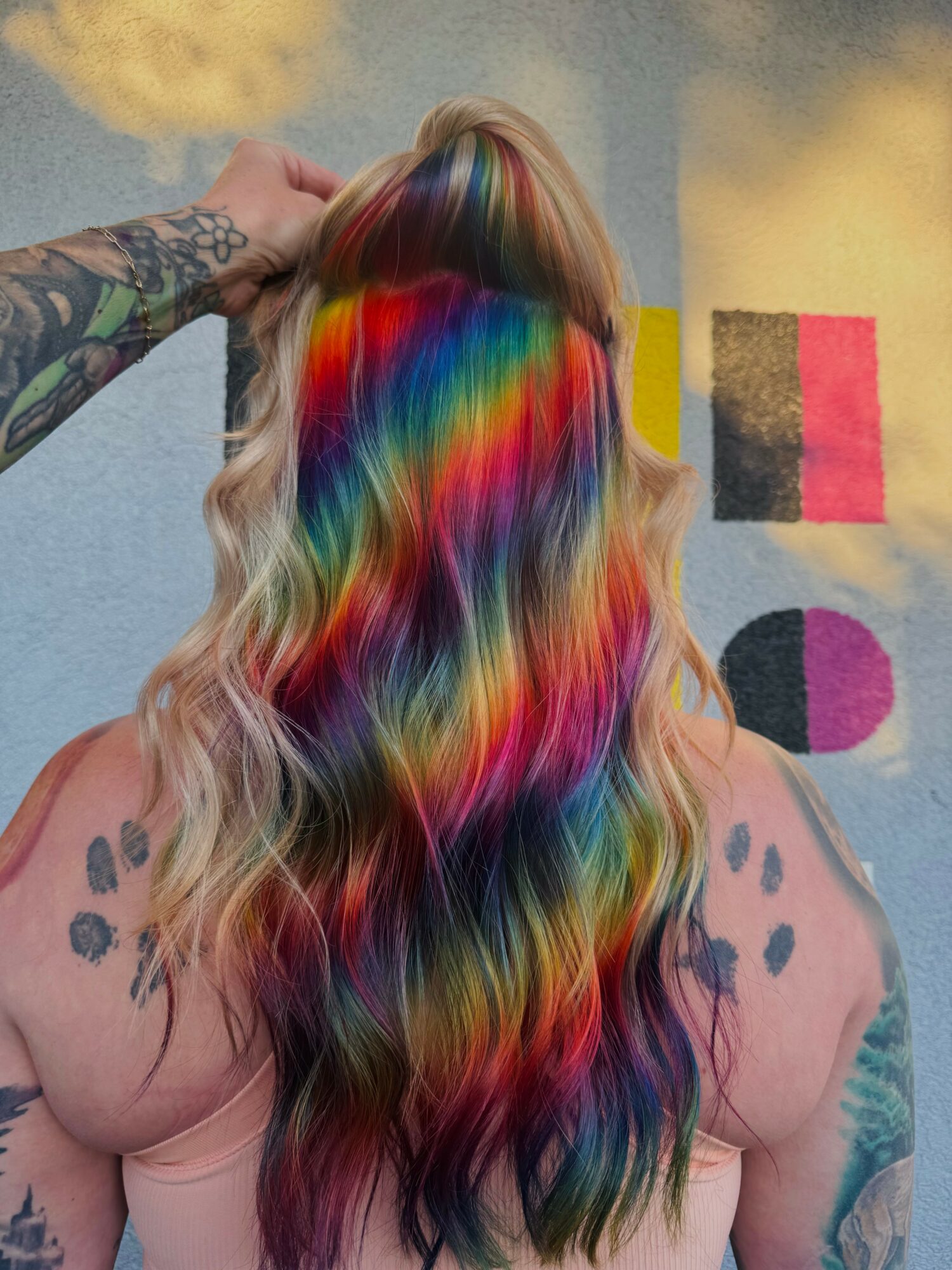 Person with long, wavy hair dyed in rainbow colors, back view, tattoos on shoulders, hand on head, colorful background.