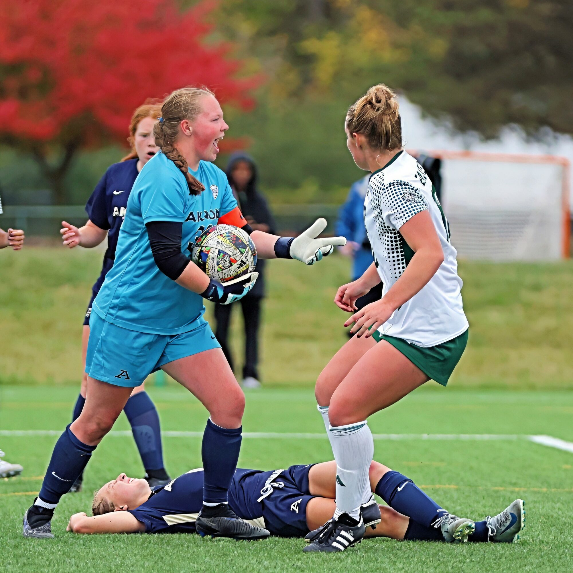 Two female soccer players, one in a blue goalkeeper uniform and the other in a white and green uniform, argue on the field, with a player lying on the ground nearby.