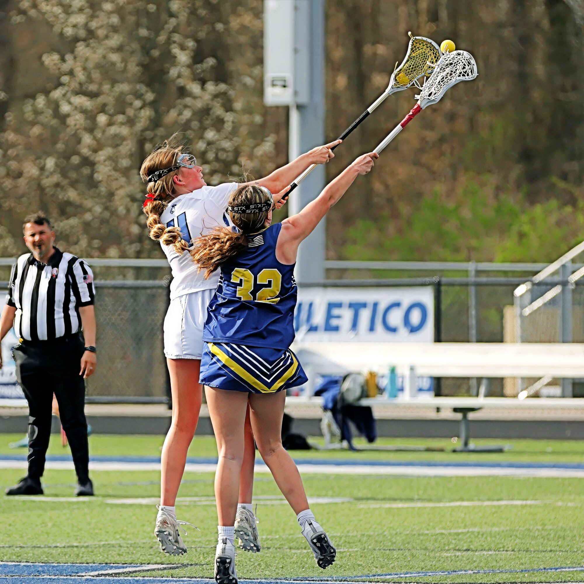 Two female lacrosse players reach for the ball with sticks during a game, with a referee and field in background.