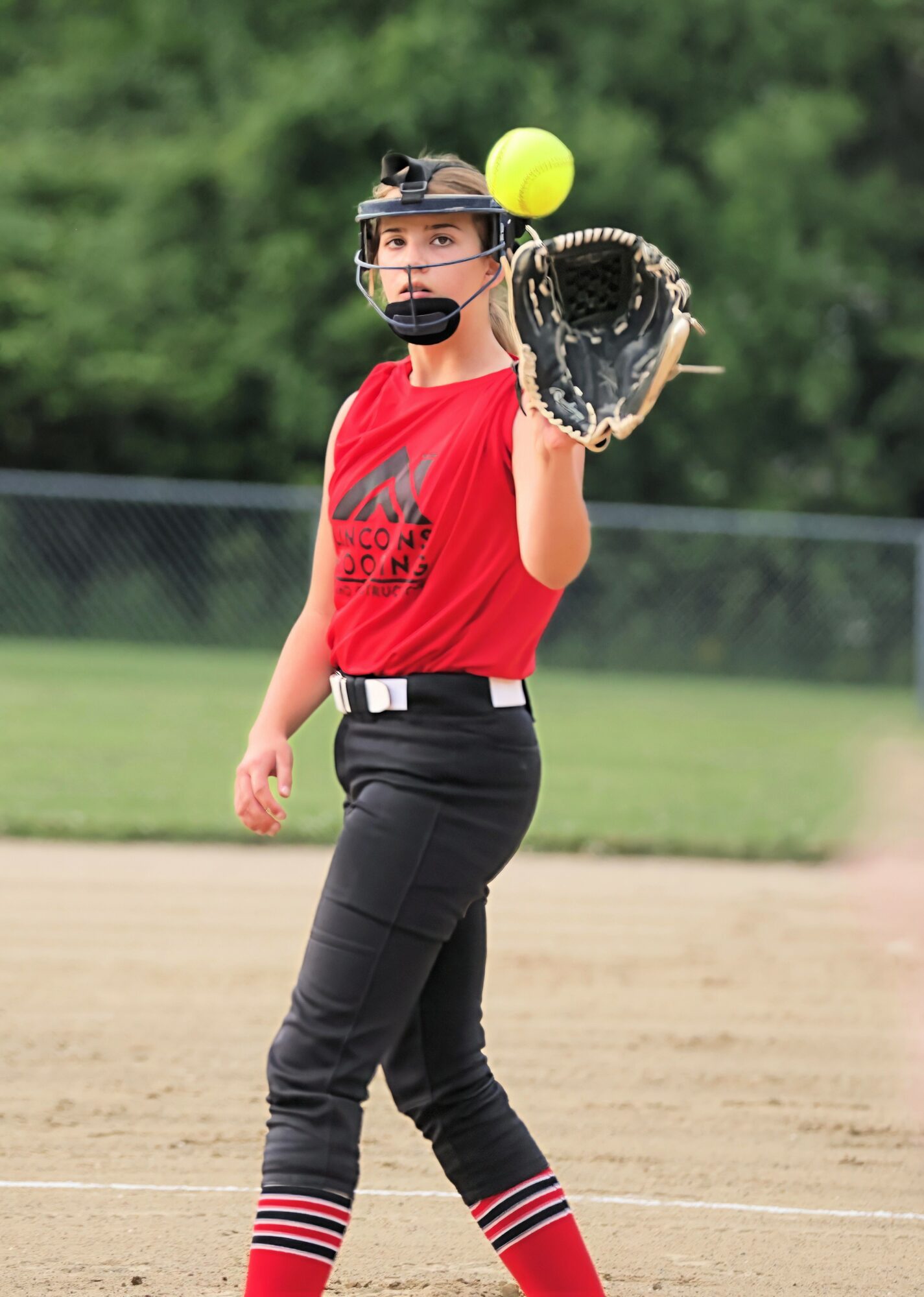 Female softball player in red shirt and black pants prepares to catch a yellow ball with her glove on a field.