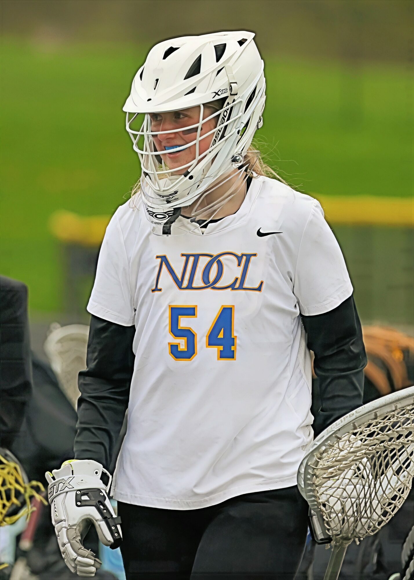 Female lacrosse player wearing a helmet and jersey with number 54, holding a lacrosse stick, outdoors on a field.