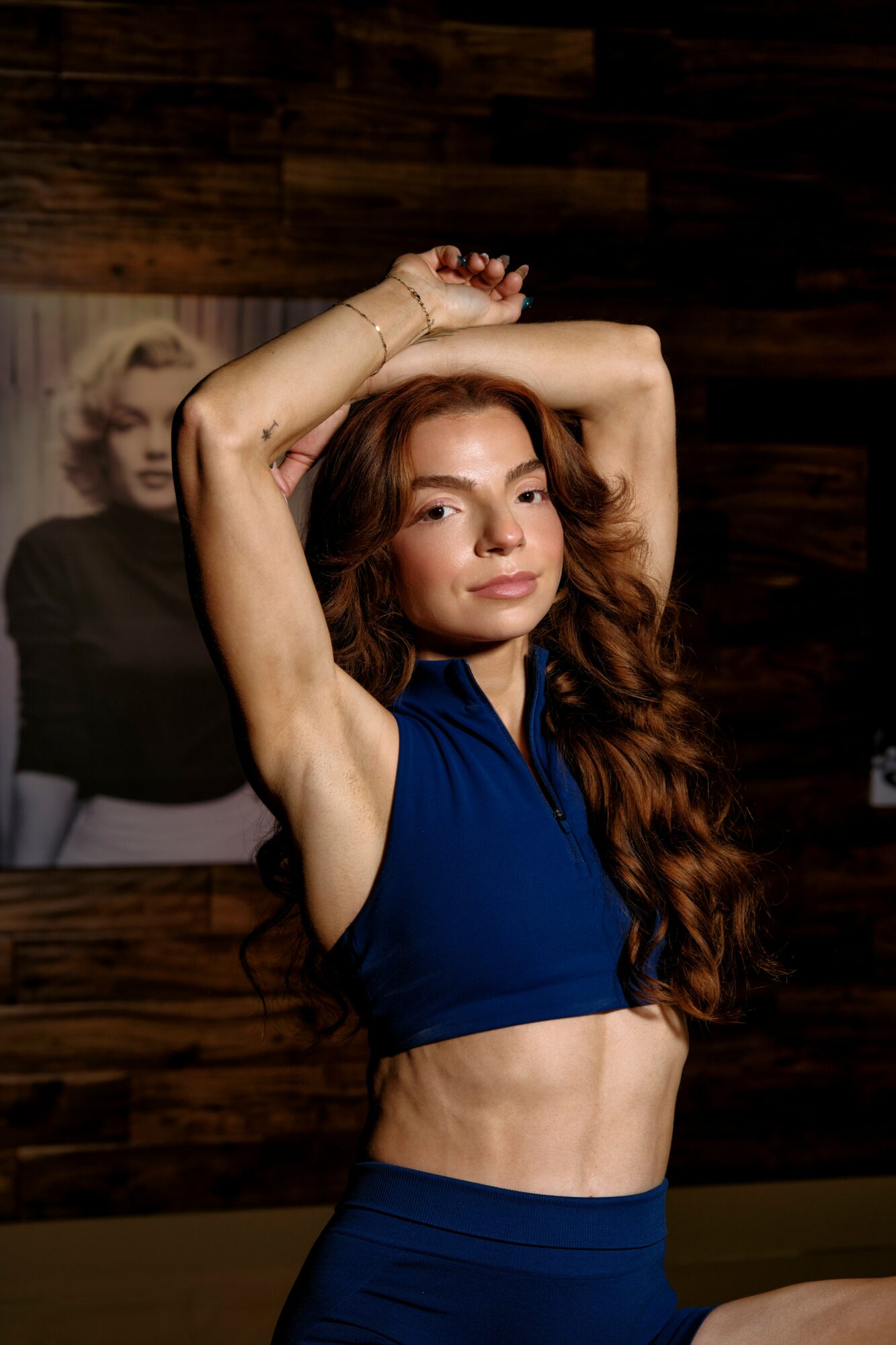 Young woman with long wavy hair in a blue sports bra and matching high-waisted pants, arms raised above head, smiling.