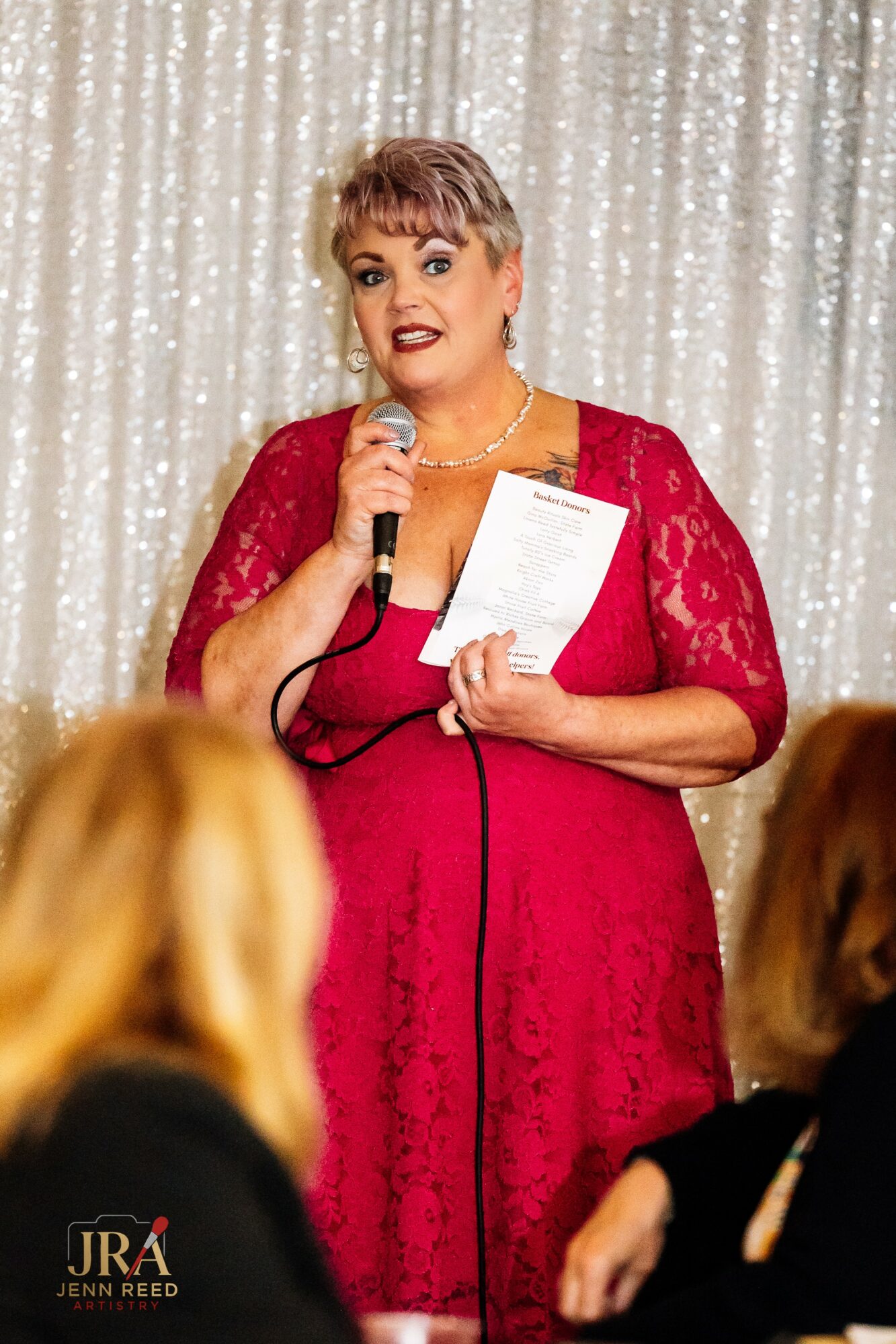 Woman with short hair in a red lace dress speaking into a microphone, holding papers, in front of a glittery backdrop.
