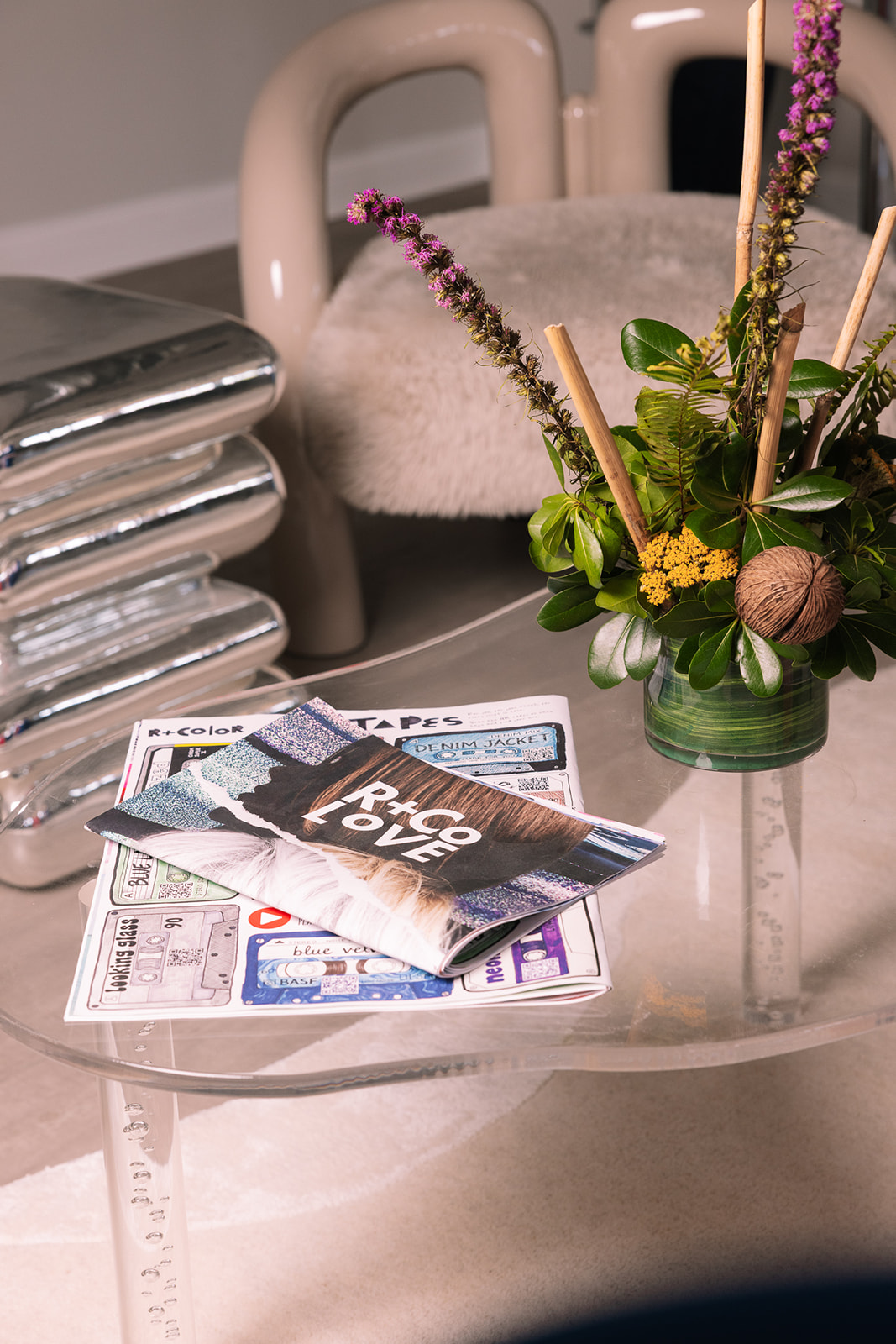 Flower arrangement with purple and yellow flowers in a green vase, magazines and a ruler on a glass table, background with stacked items.