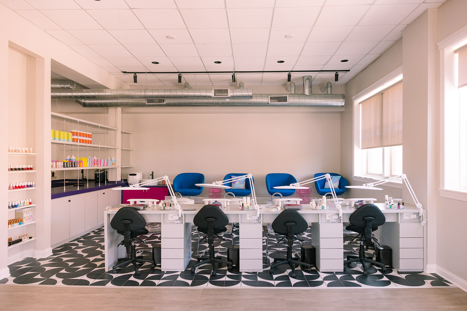 Nail salon with manicure stations, chairs, and shelves with nail polish bottles, bright natural light, patterned floor, and white walls.