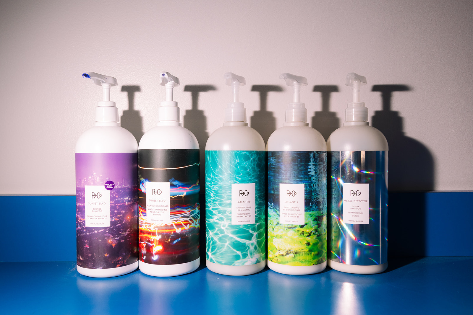Five bottles with pump dispensers lined up on a blue surface, with shadows cast on a plain background.