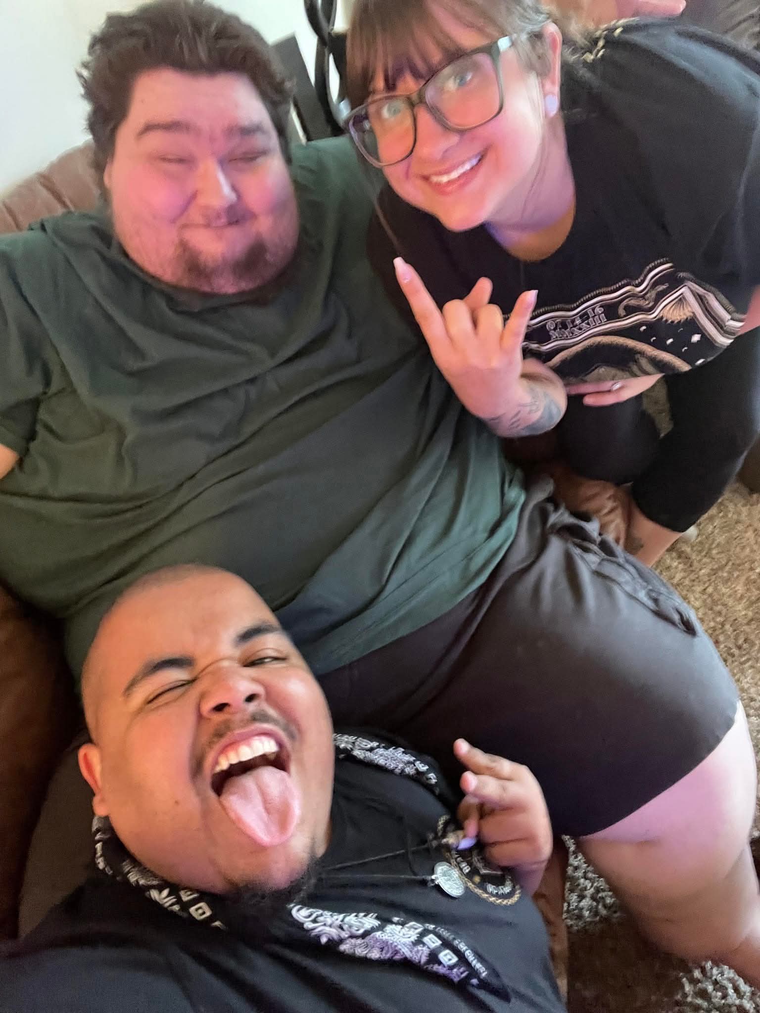Three people smiling and making gestures indoors, one lying down, two kneeling, casual clothing, cheerful expressions.