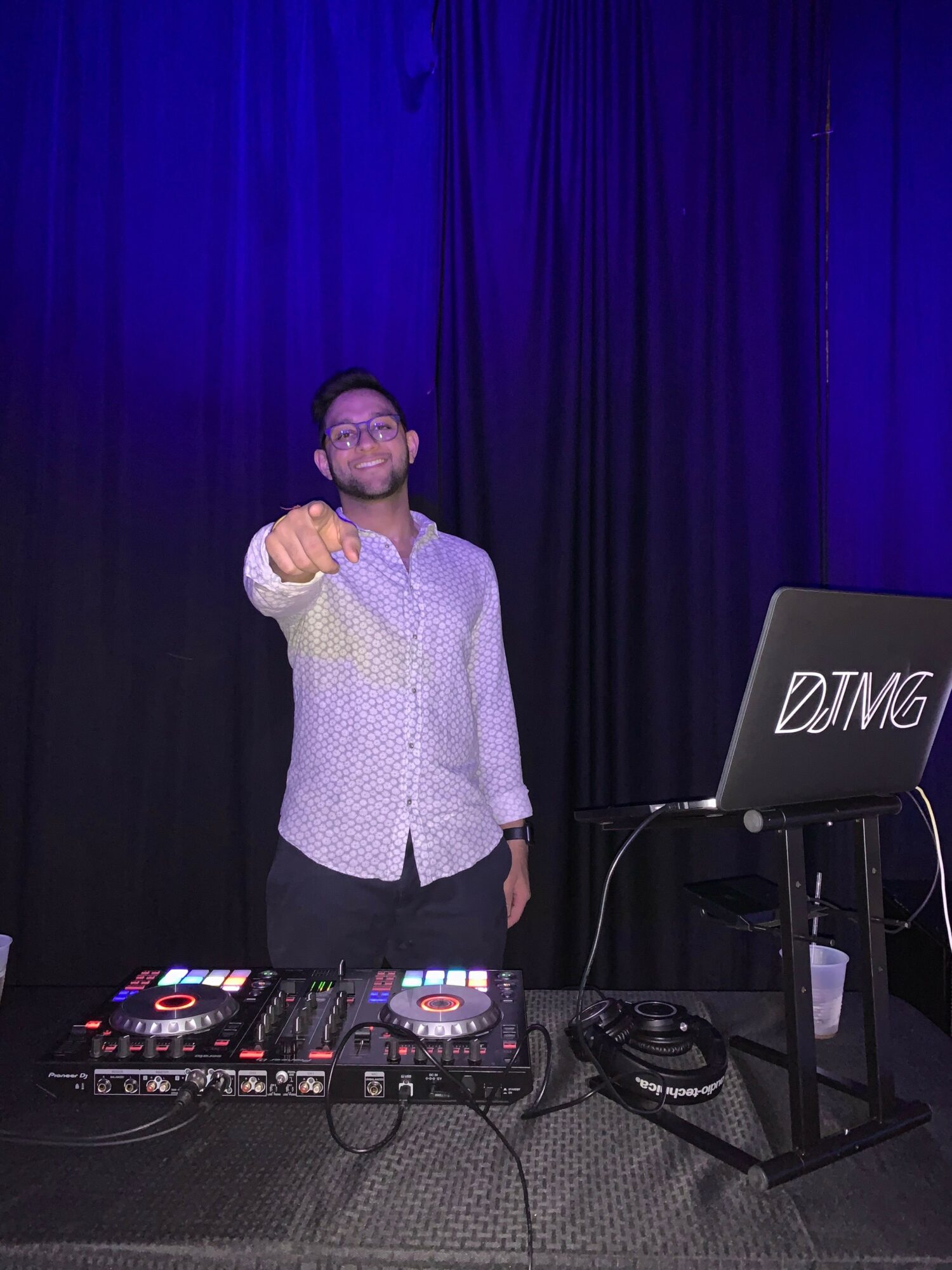Person standing on stage with DJ equipment, giving a thumbs-up, in front of a dark curtain backdrop.