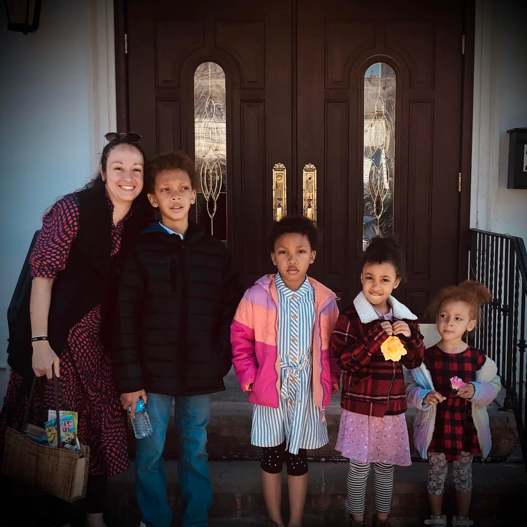 Group of five people, including a woman and four children, standing in front of a wooden door, smiling outdoors.