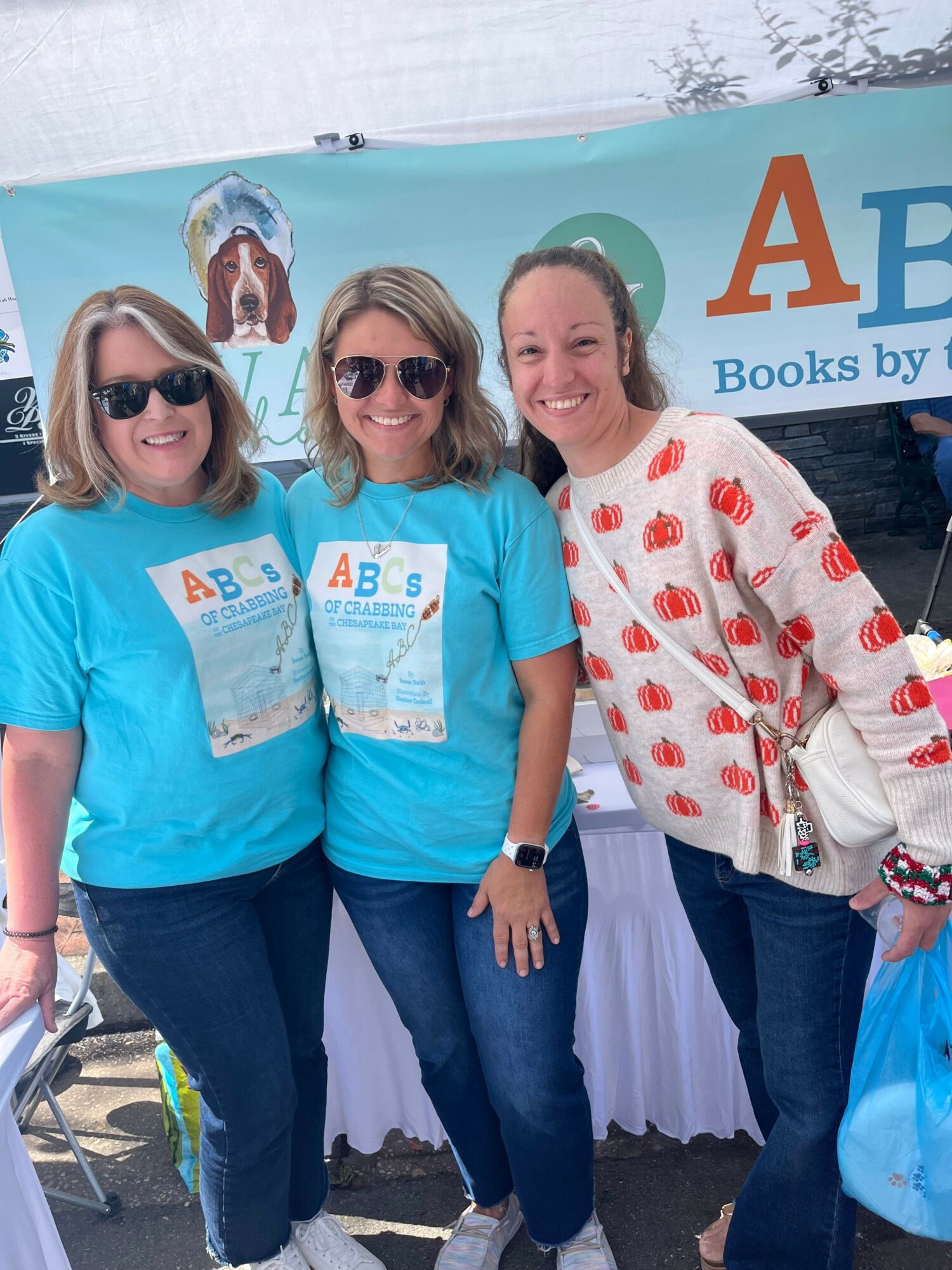 Three women standing outdoors in front of a banner, smiling, wearing sunglasses and casual clothing, with a dog illustration on the banner.