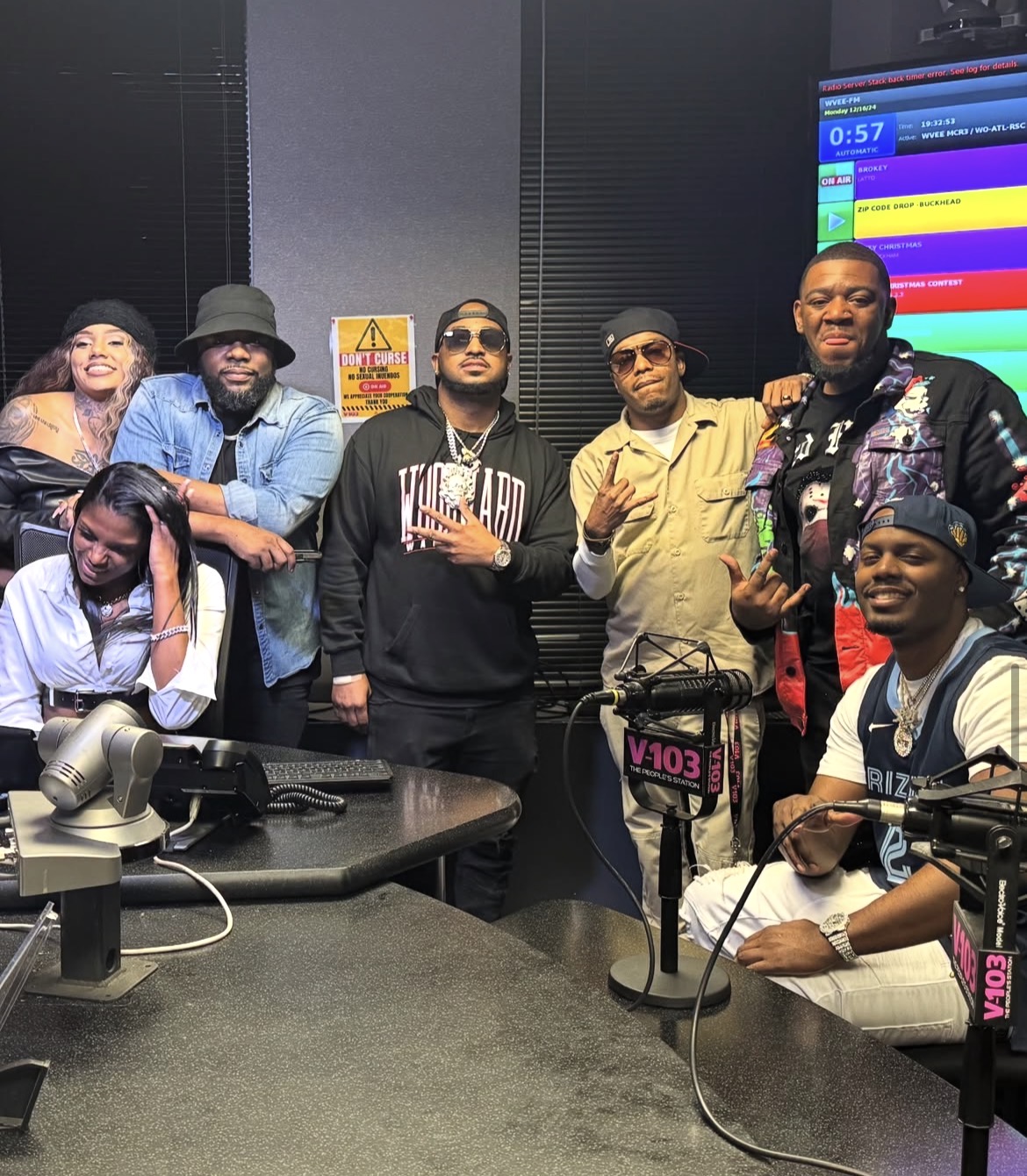 Group of seven people in a radio studio, some wearing headphones, with a colorful screen in the background.