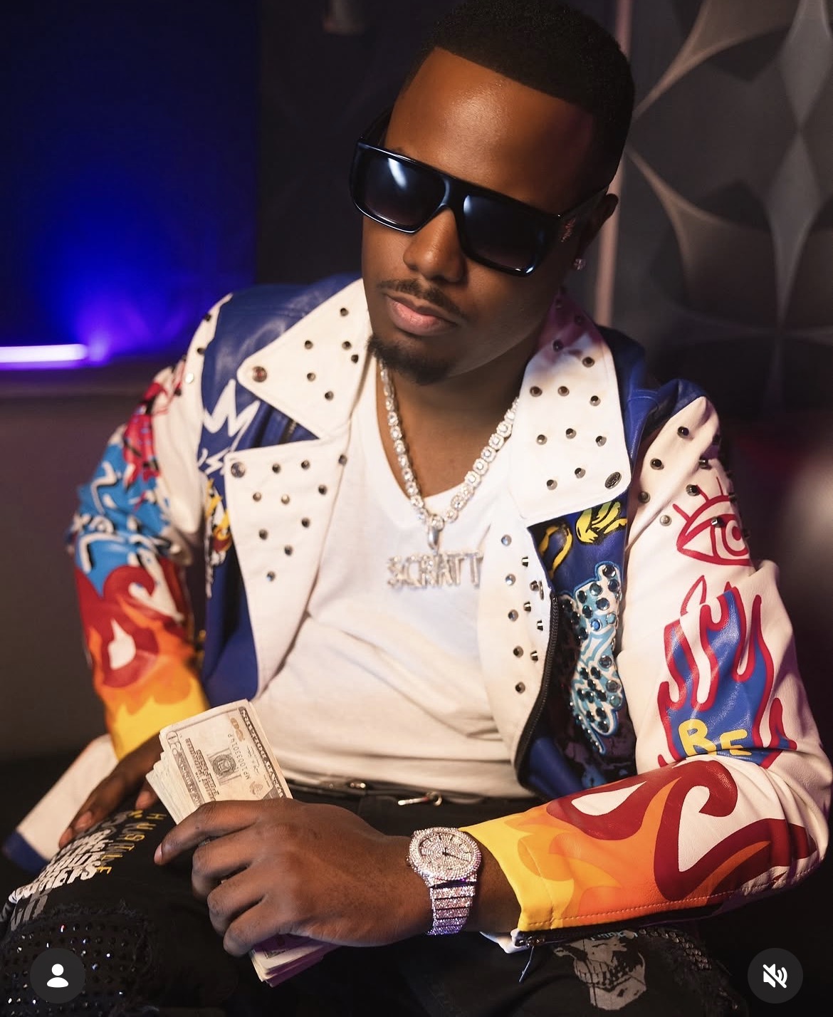 Man wearing sunglasses and a colorful jacket, holding money, in a dimly lit setting.