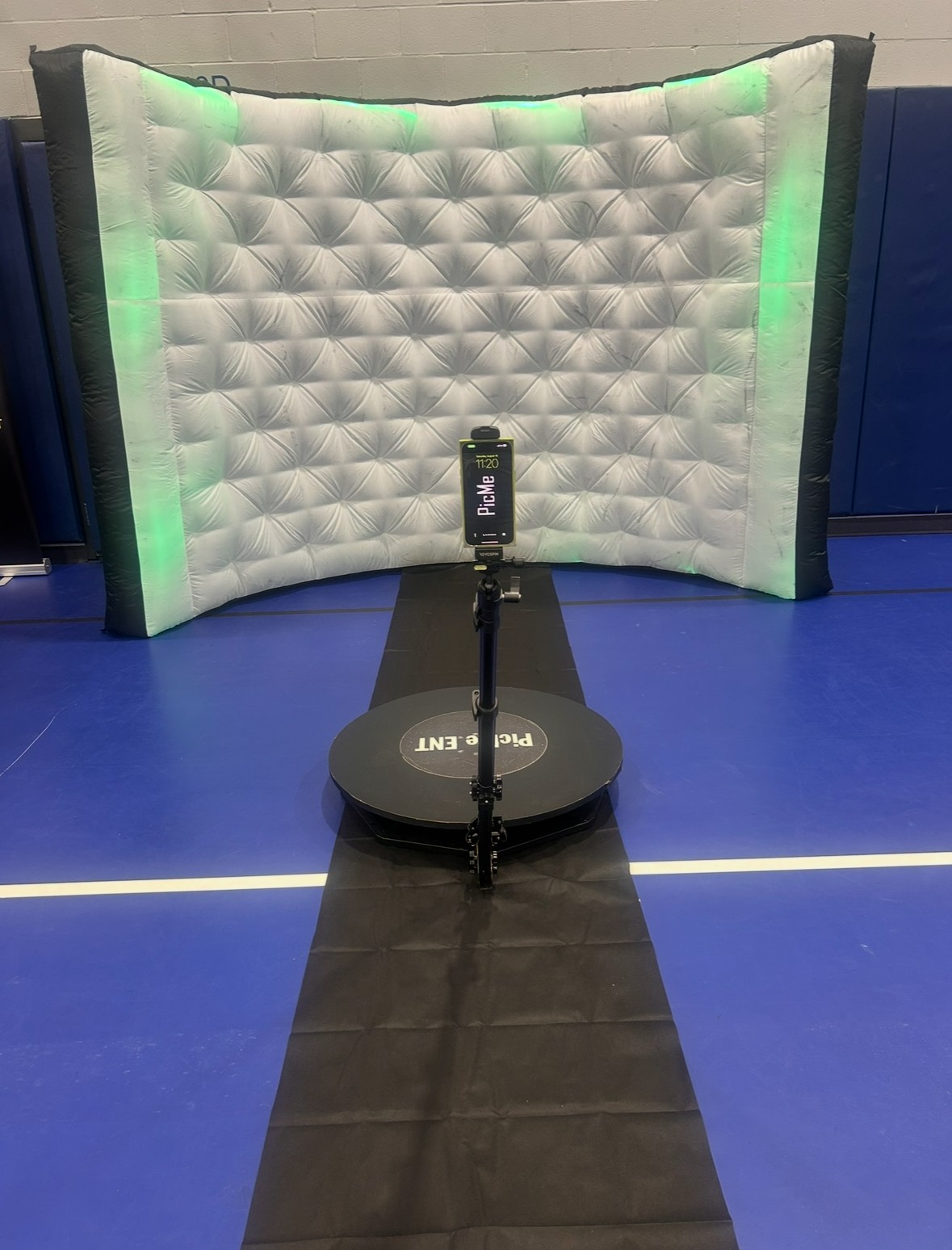 A white padded wall with green lighting, a microphone on a stand, and a black circular base on a blue floor.