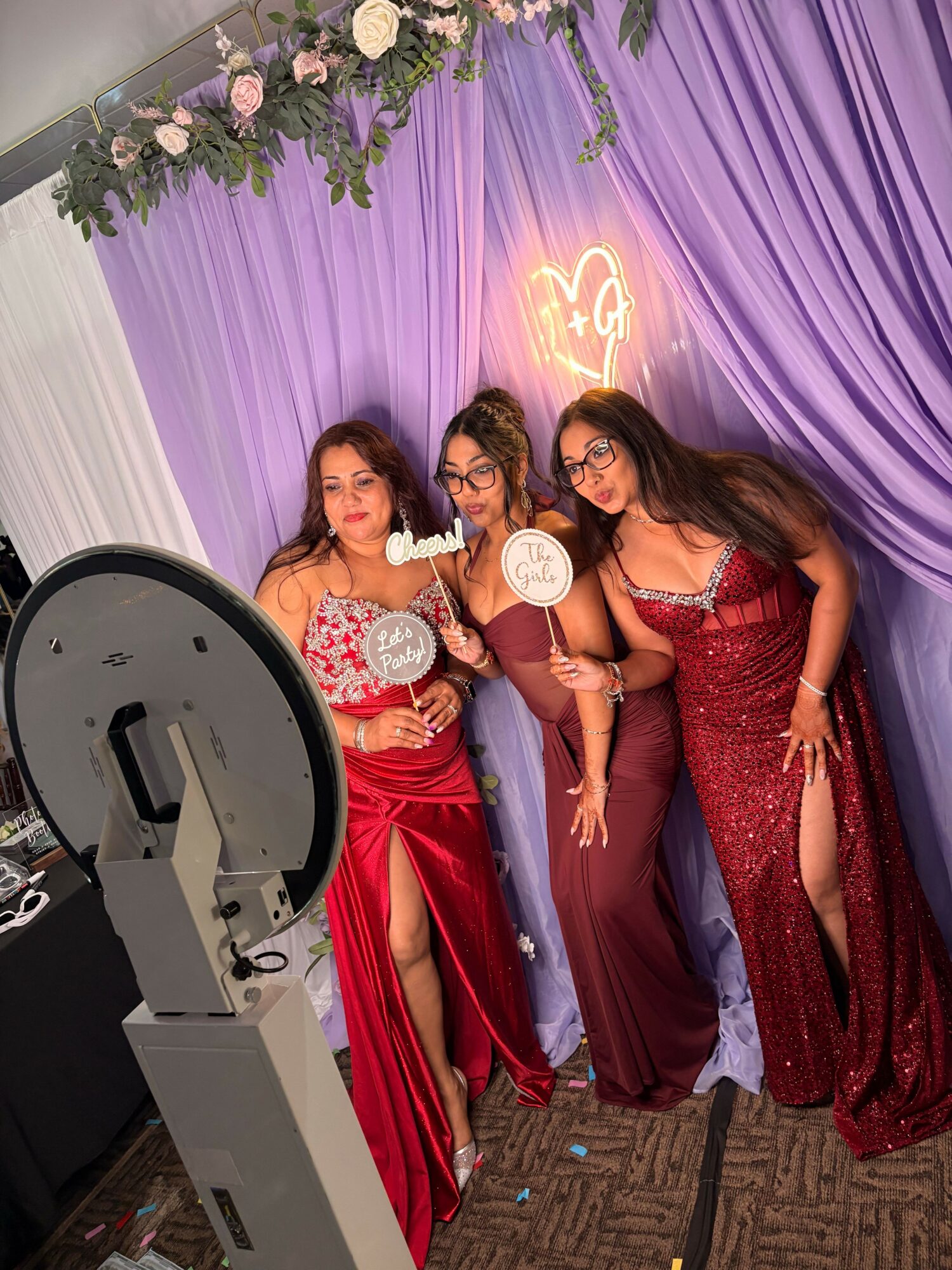 Three women in red dresses pose together in front of purple and white drapery with floral decorations, holding round signs.