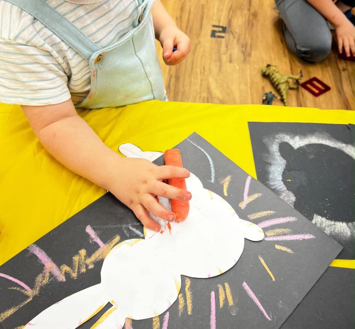 Child using orange chalk to draw on black paper, with other drawings and toys nearby, on a yellow surface.