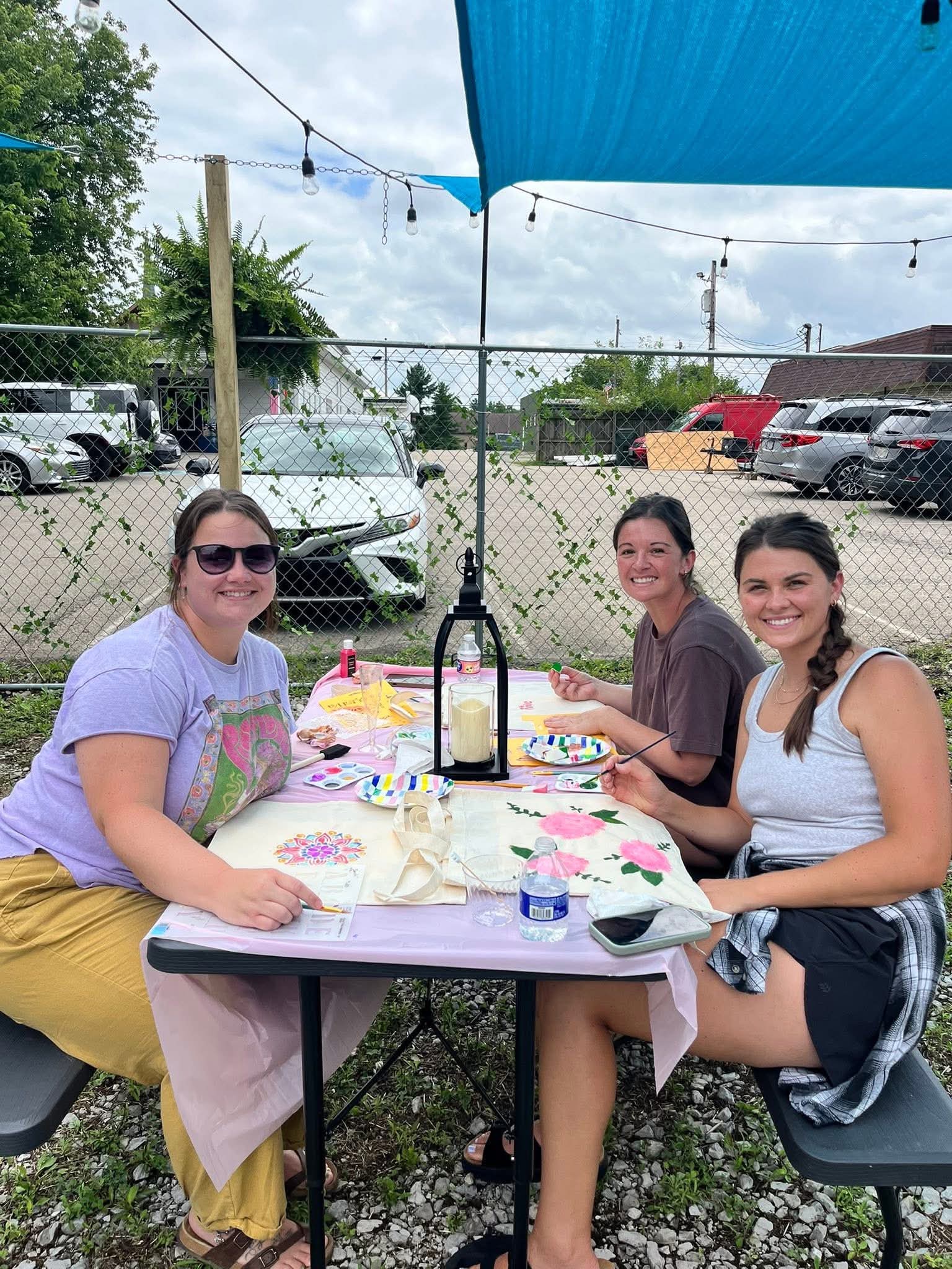 Three women sit at a table outdoors under a blue canopy, enjoying a meal with plates, drinks, and decorations.