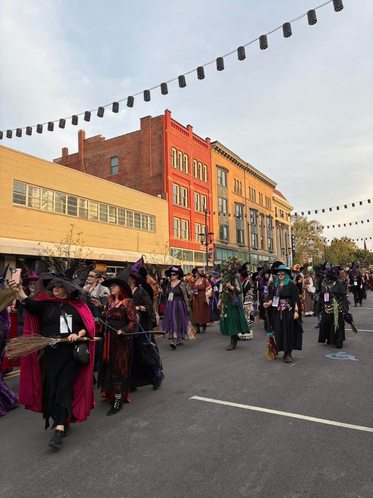 People gather on a street with historic buildings, some holding umbrellas, under string lights, during daytime.