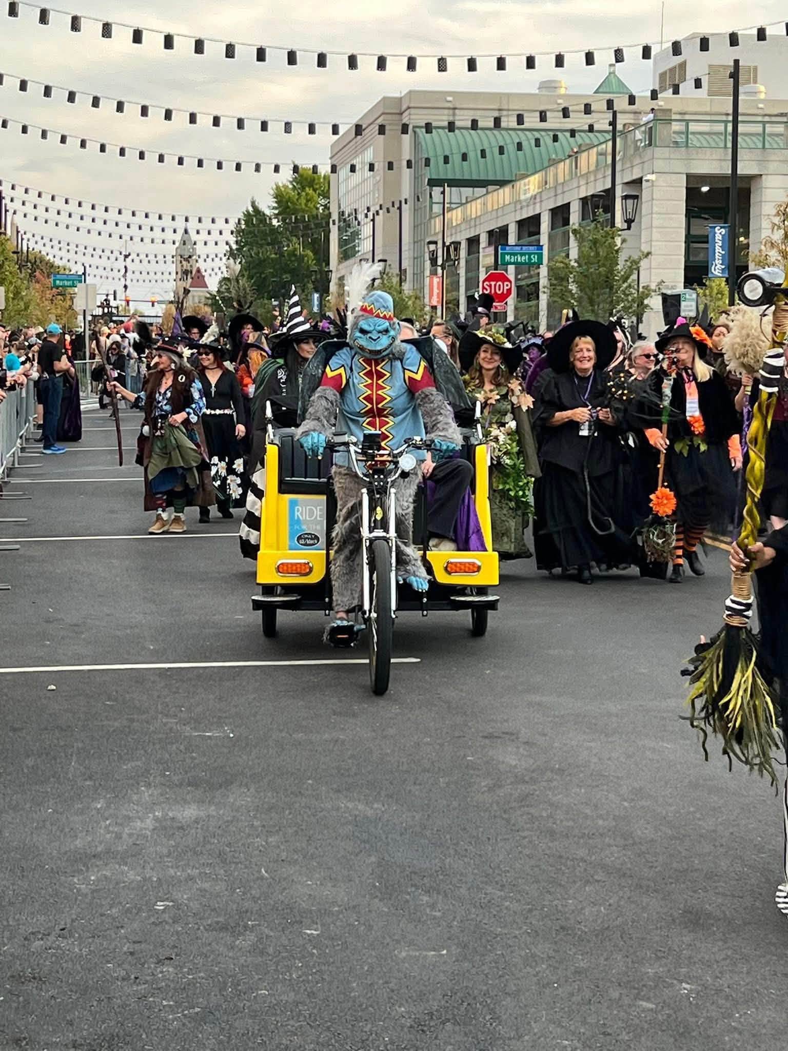 Parade with people in costumes walking down a street decorated with hanging banners, some holding broomsticks, with buildings in background.