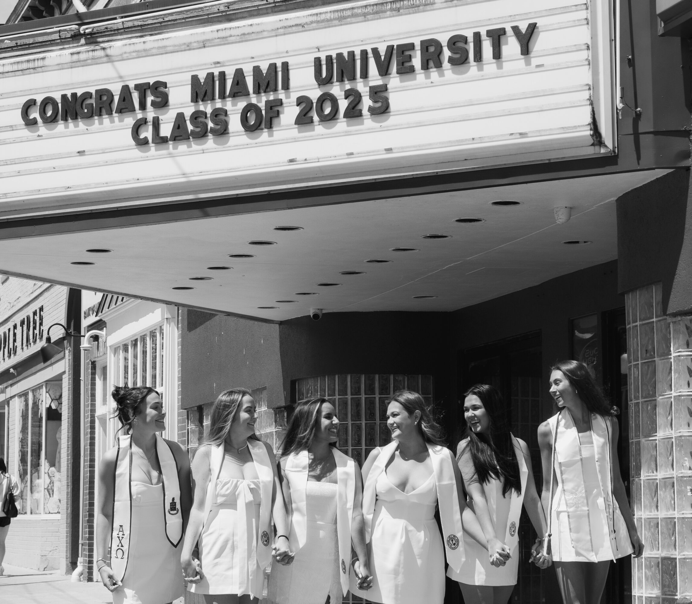 Group of seven women in white dresses standing outside a building with a congratulatory sign for Miami University, Class of 2025.