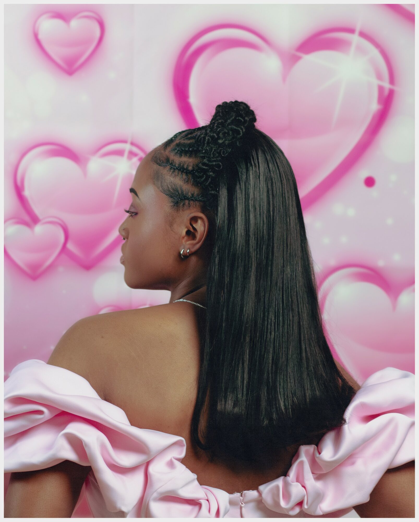 Side profile of a woman with long, straight black hair and a braided updo, wearing a pink ruffled dress, against a pink heart background.