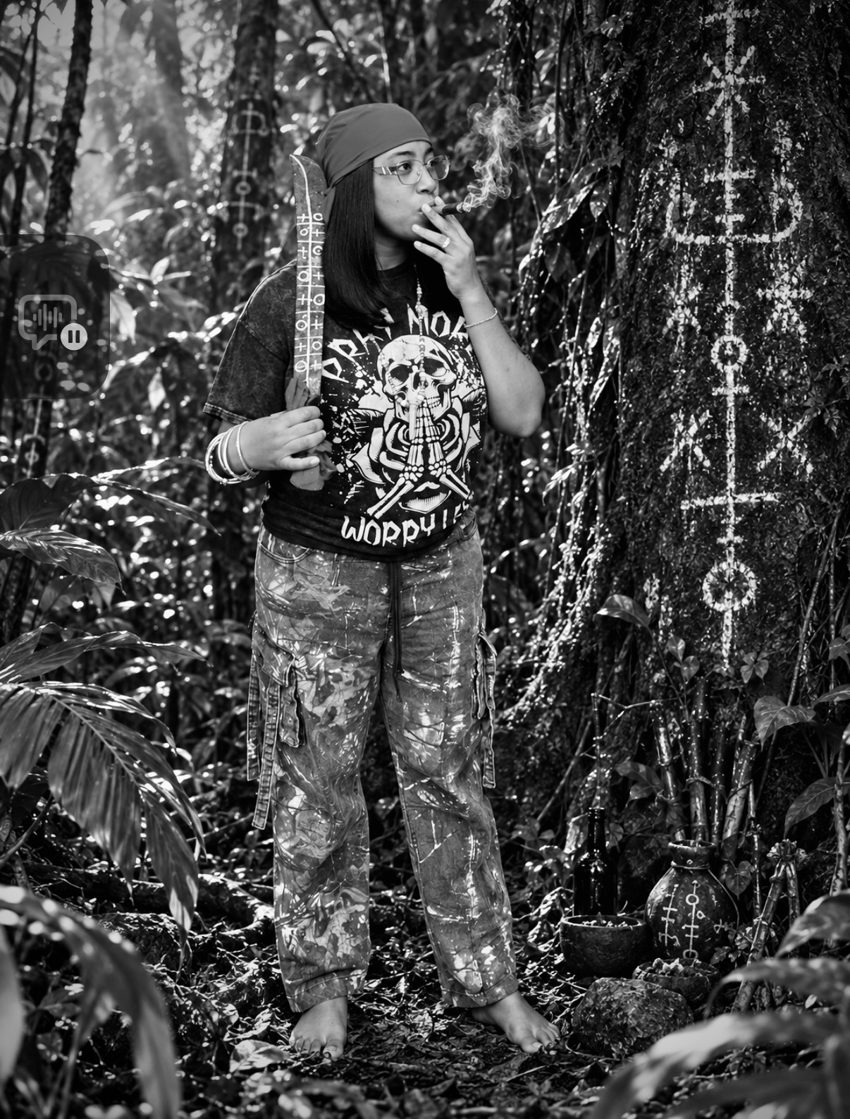 Person standing in a forest with trees and symbols on a tree trunk, wearing a bandana, glasses, and casual clothing.