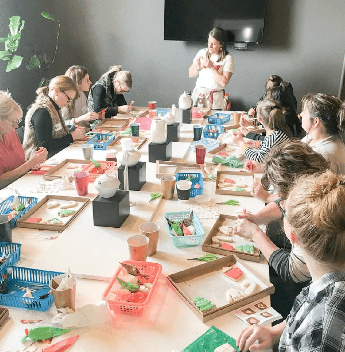 Group of people sitting around a table, a woman standing and speaking, various craft supplies on the table.