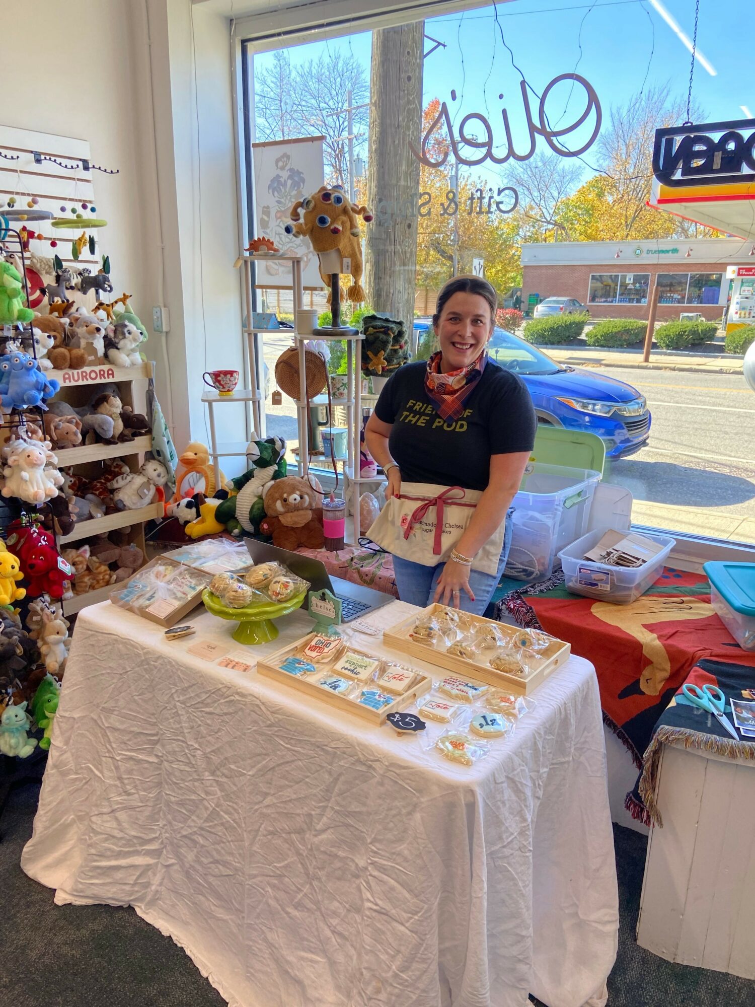Woman standing behind a table with various items, inside a store with large windows and a sign outside. Word 'Olio' visible.
