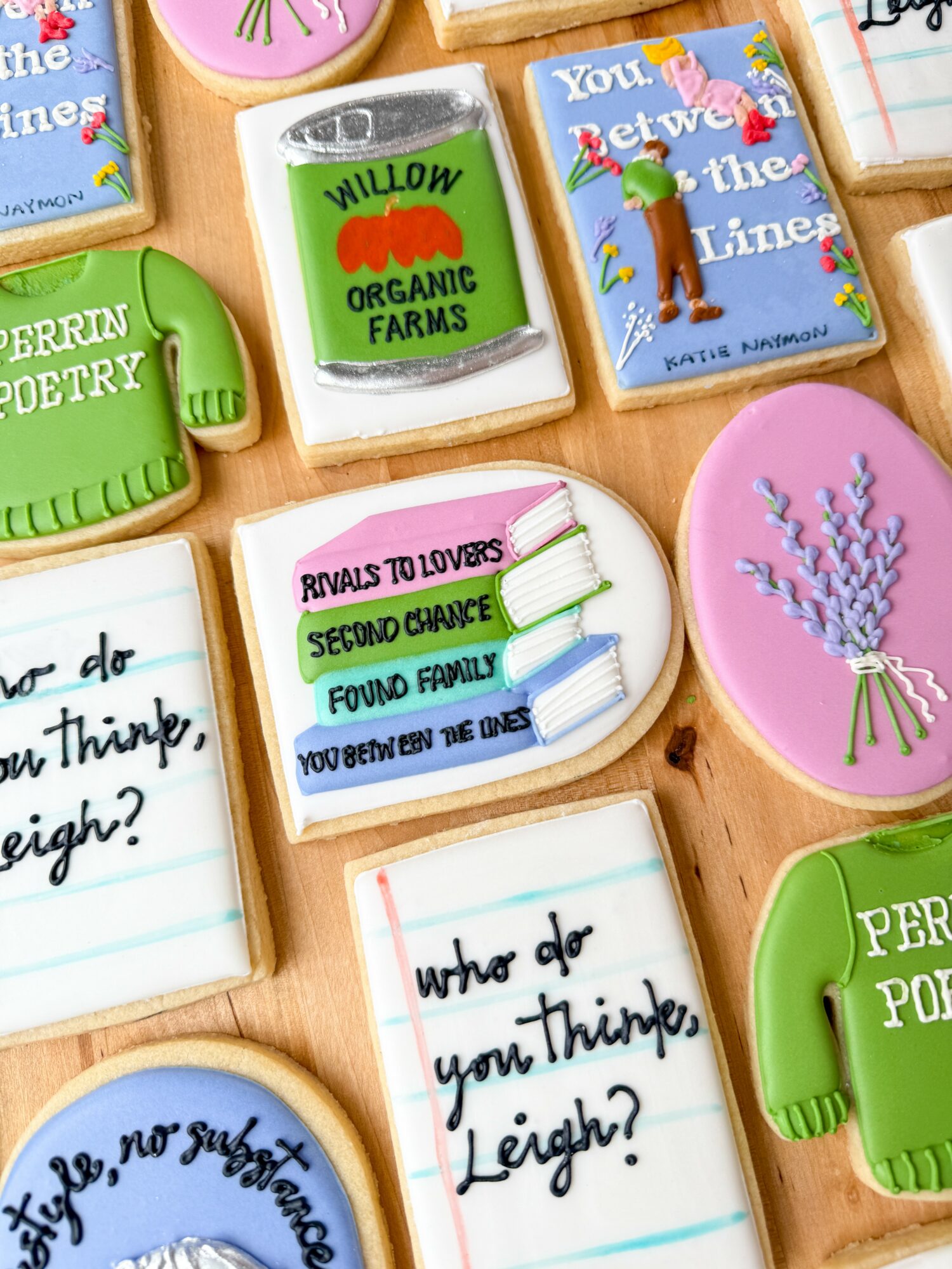 Assorted decorated cookies with colorful designs and text, including a green mug, a can, a book, and a lavender bouquet, on a wooden surface.