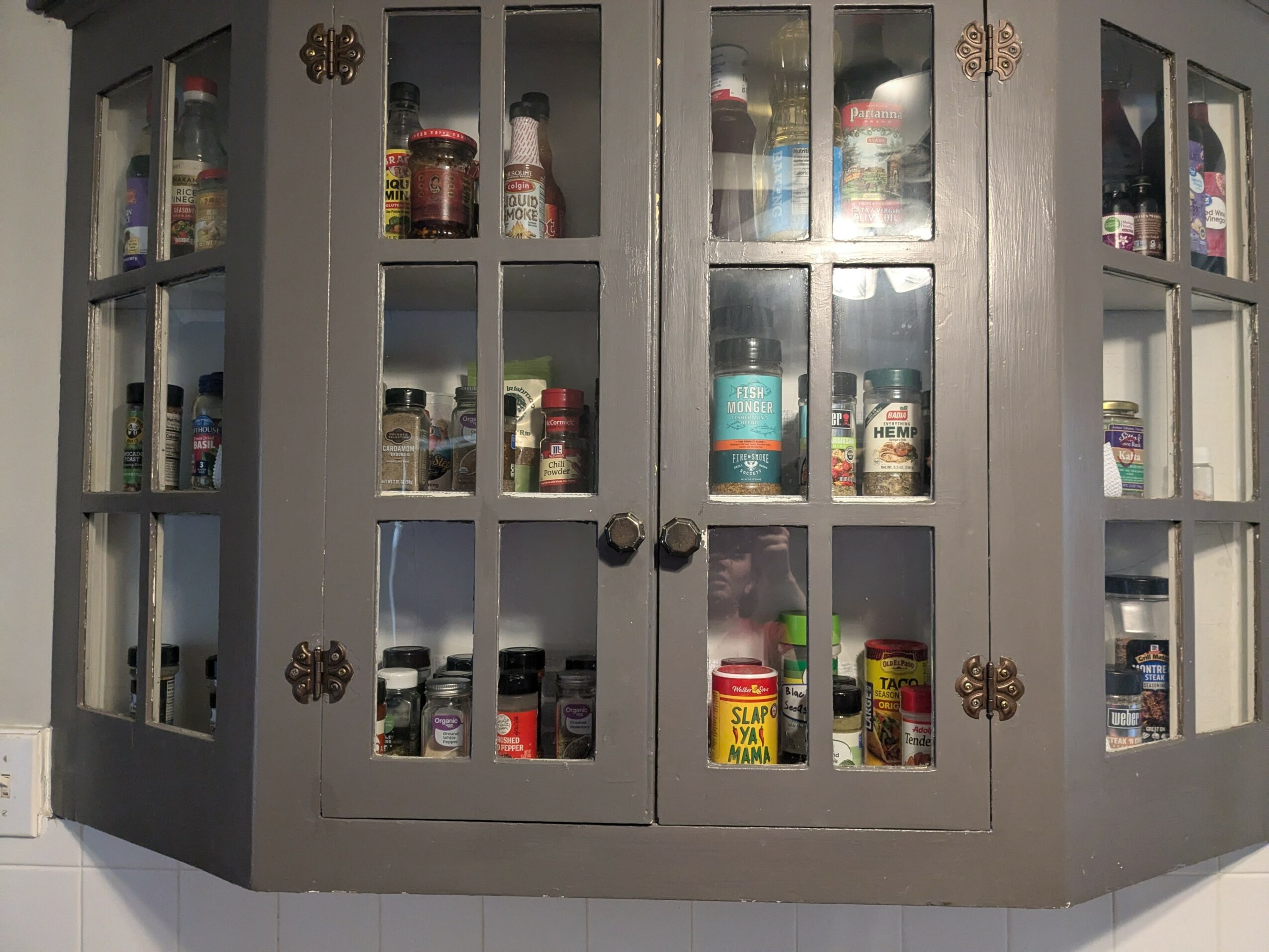 Gray kitchen cabinet with glass doors filled with various jars and containers, some with labels visible.