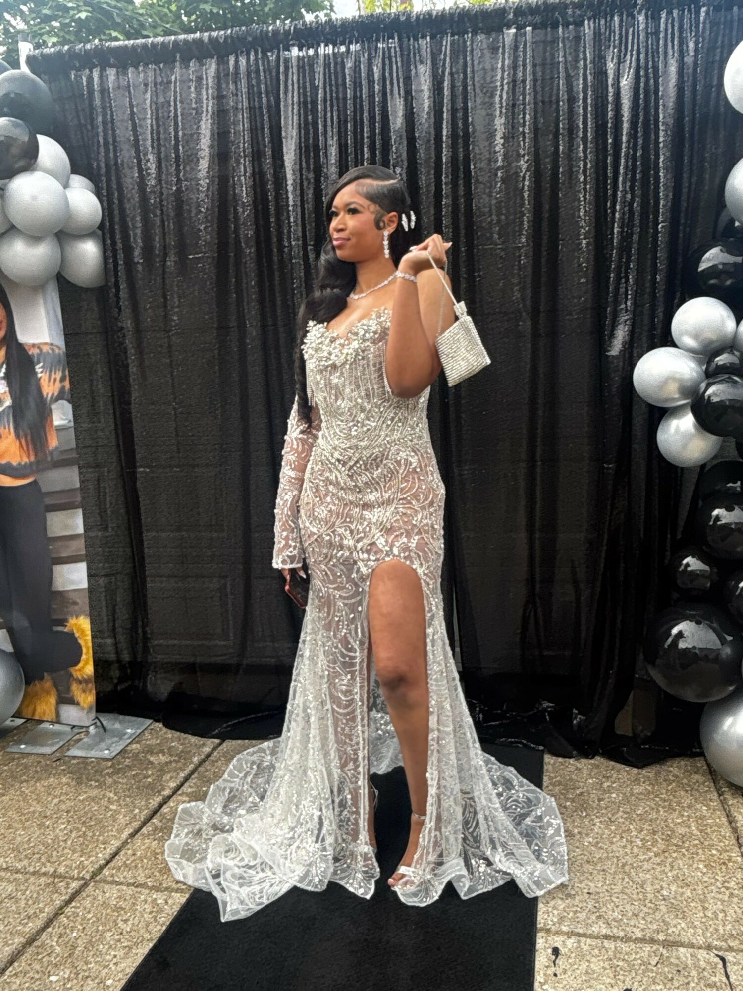 Woman in a long, sparkly gown standing in front of black curtains with balloons, talking on a phone, facing left.