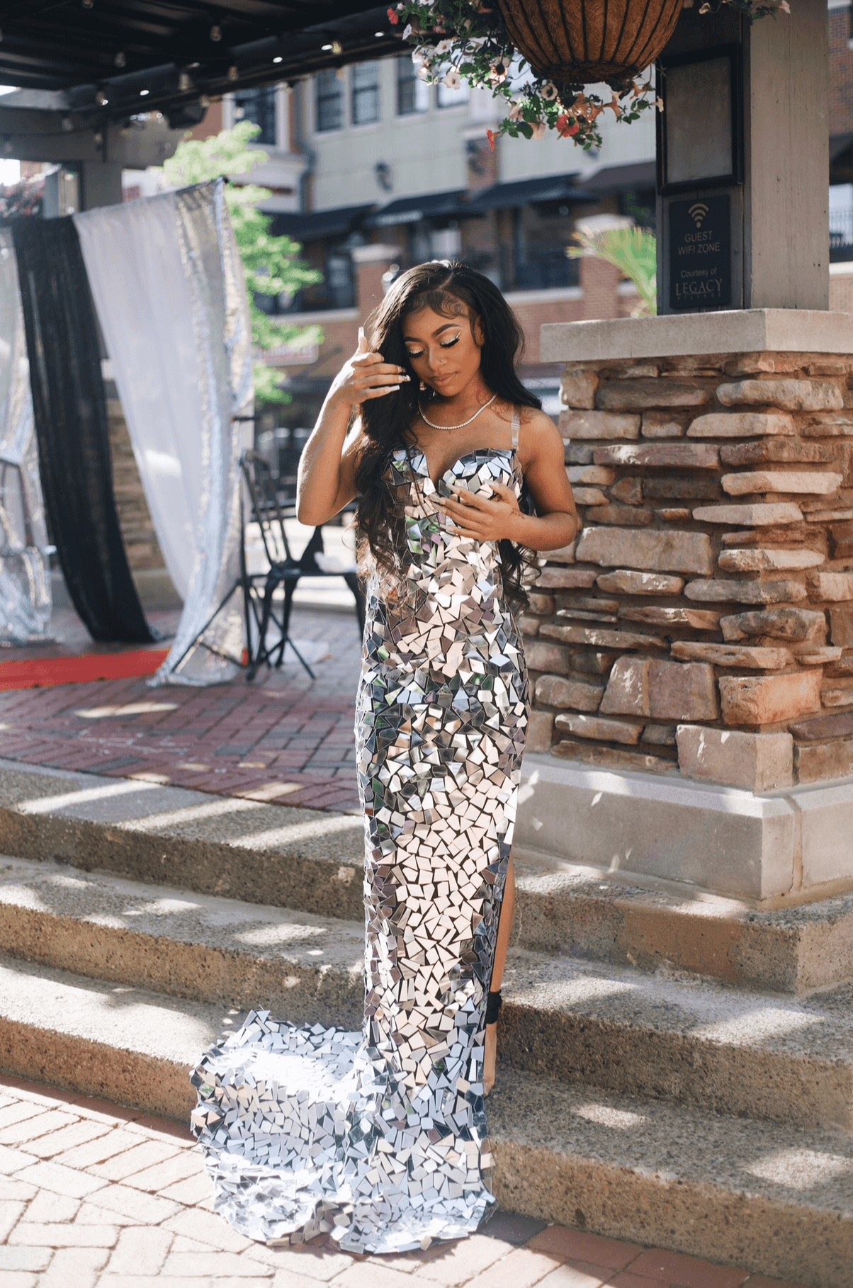 Woman in a shiny, sequined gown standing on steps outside, adjusting her hair, with a brick wall and outdoor setting behind her.