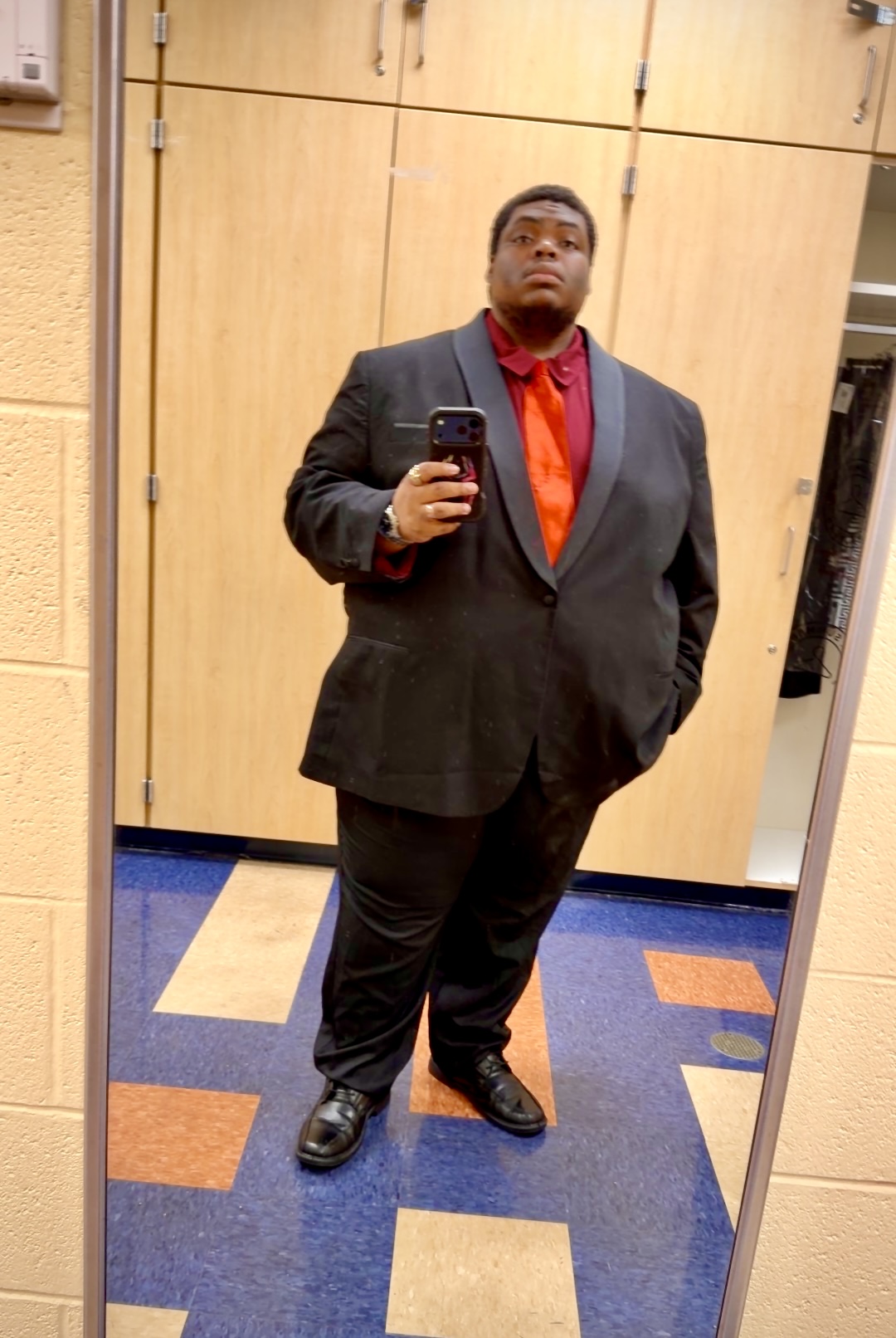 Person in a suit taking a mirror selfie in a room with wooden cabinets and colorful carpet.