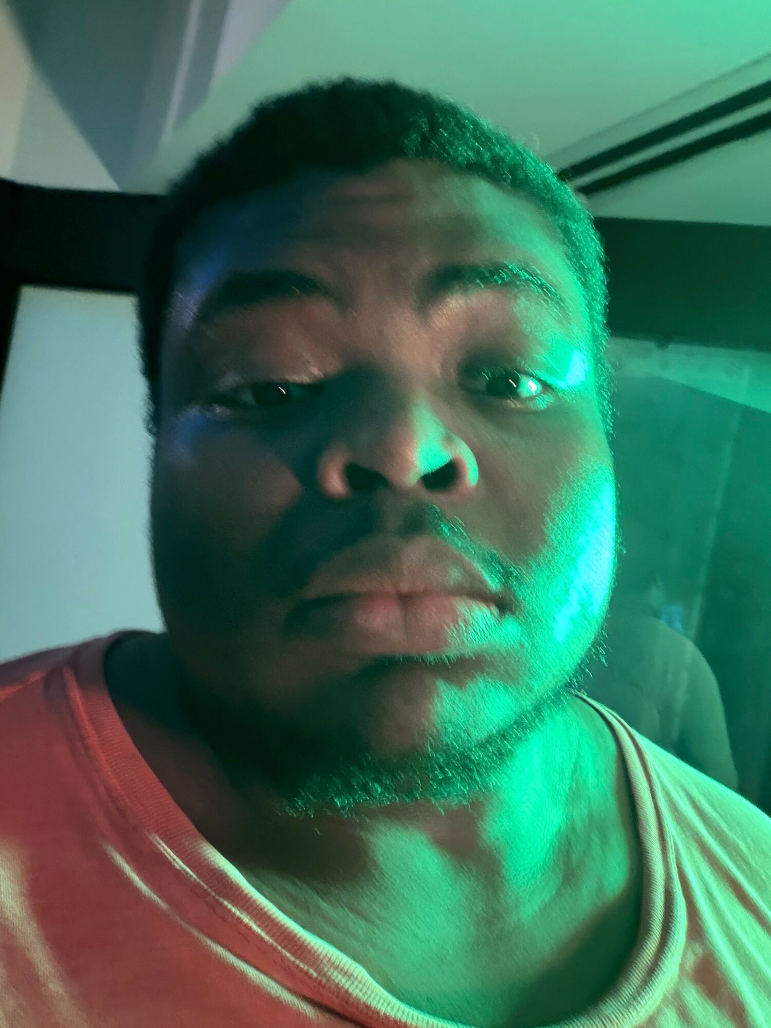 Close-up of a person with short hair and a beard, illuminated by green light, wearing a red shirt.