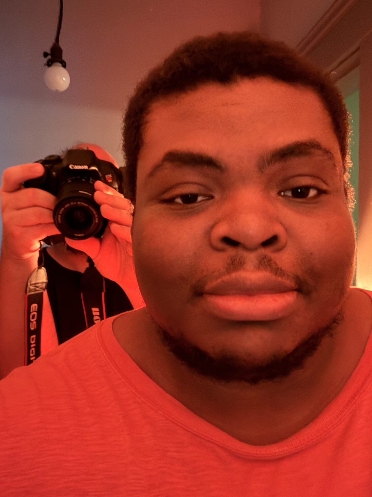 Young man with short curly hair and a beard, wearing a red shirt, looking at the camera, with a person taking a photo in the background.