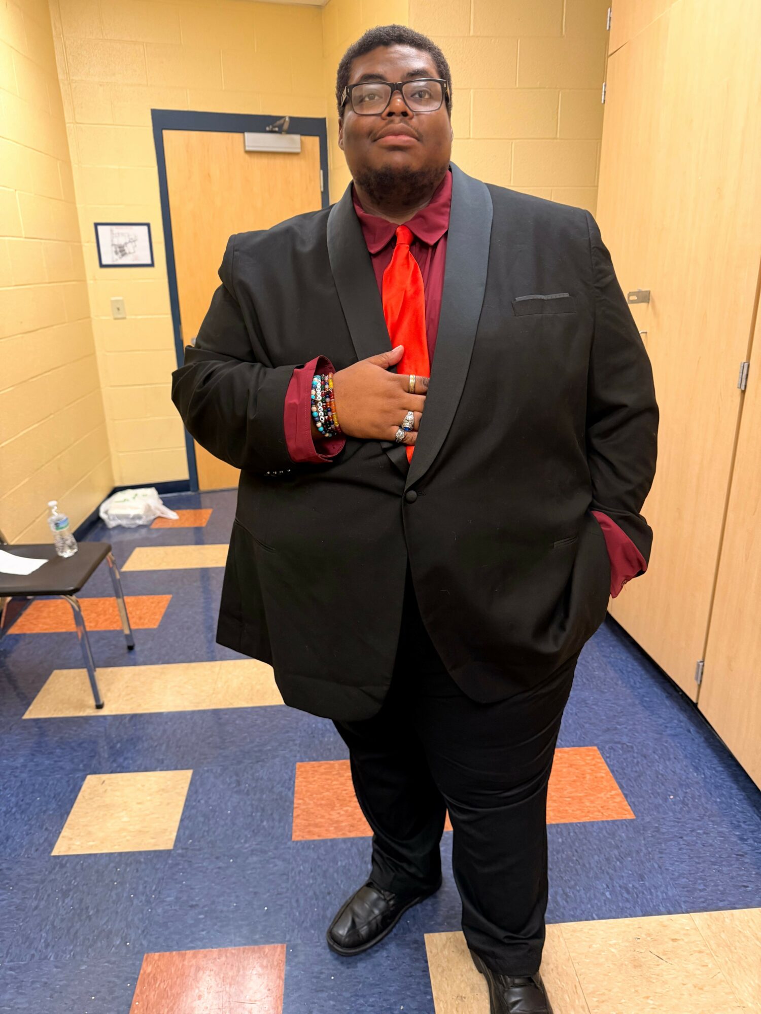 Man standing indoors on a blue and orange carpet, wearing a black jacket, red shirt, and black pants, holding a phone.