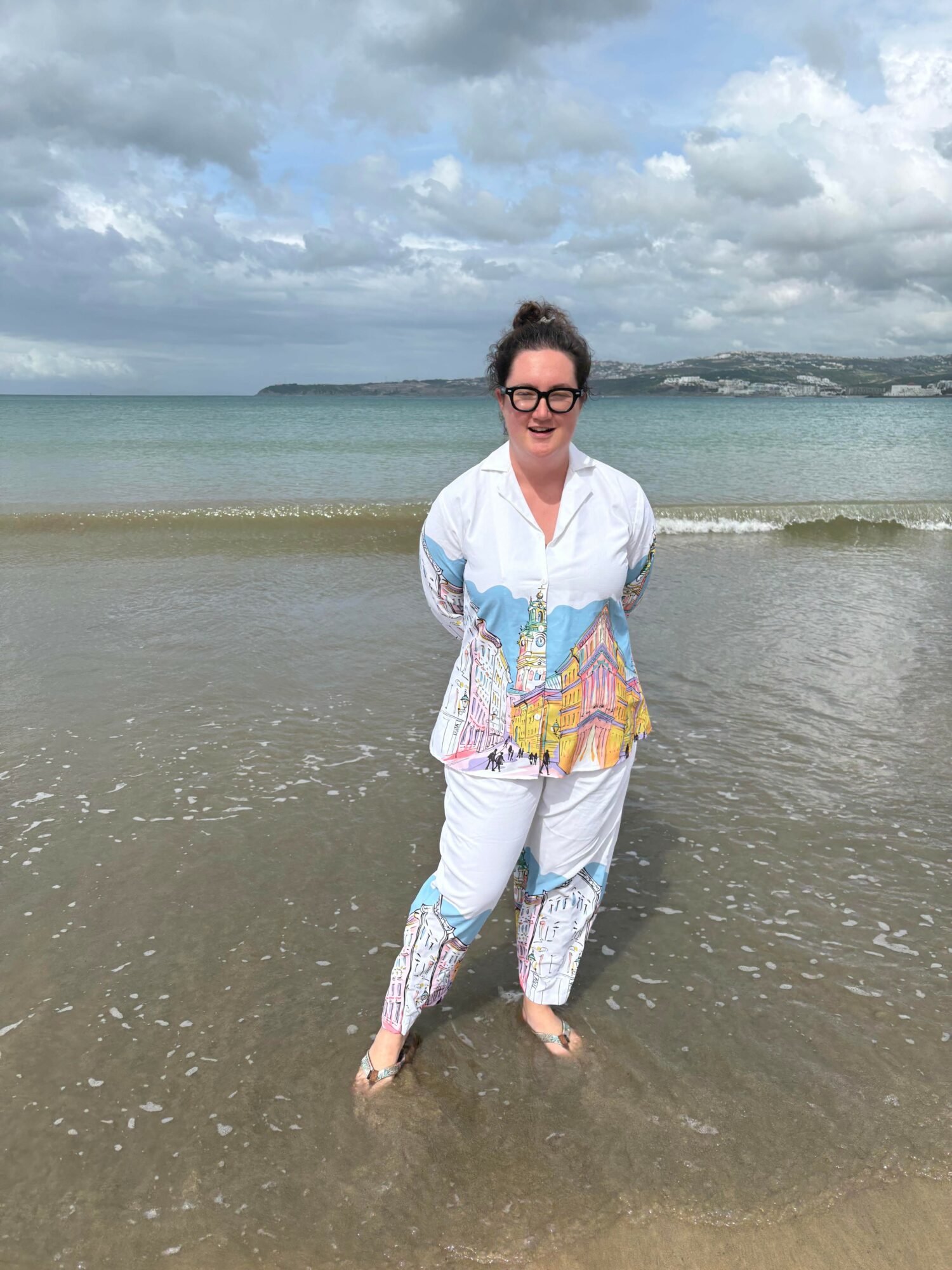Woman standing in shallow water at the beach, wearing a colorful outfit, with ocean and cloudy sky in background.