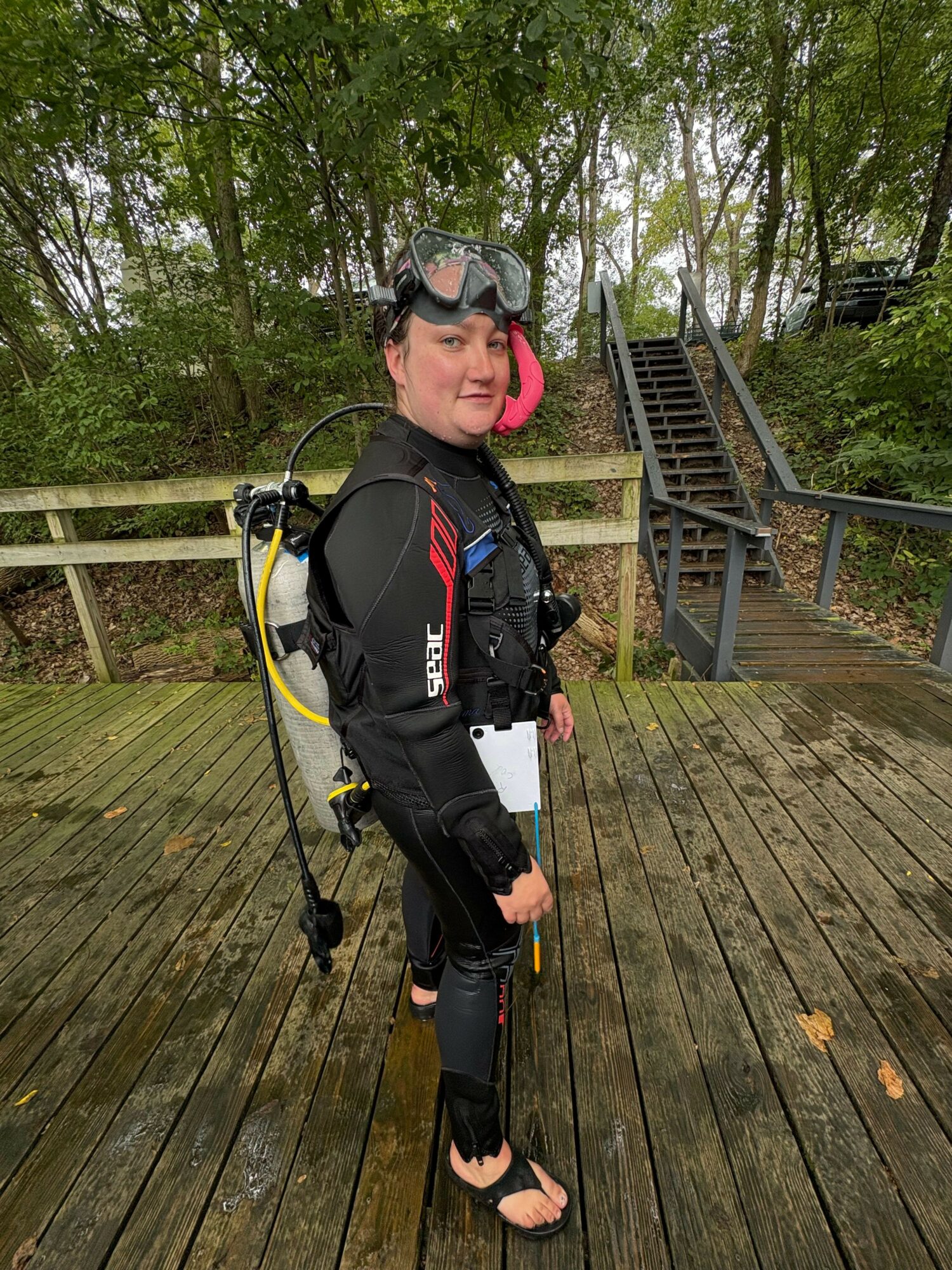 Person in black wetsuit with scuba gear standing on wooden deck surrounded by trees.