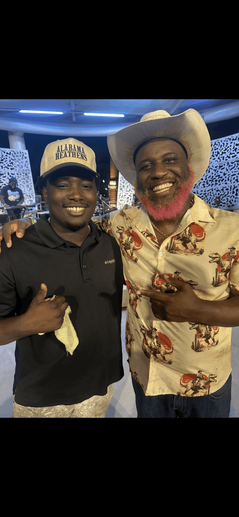Two men smiling, one wearing a cowboy hat and floral shirt, the other in a cap and dark shirt, in a decorated indoor setting.