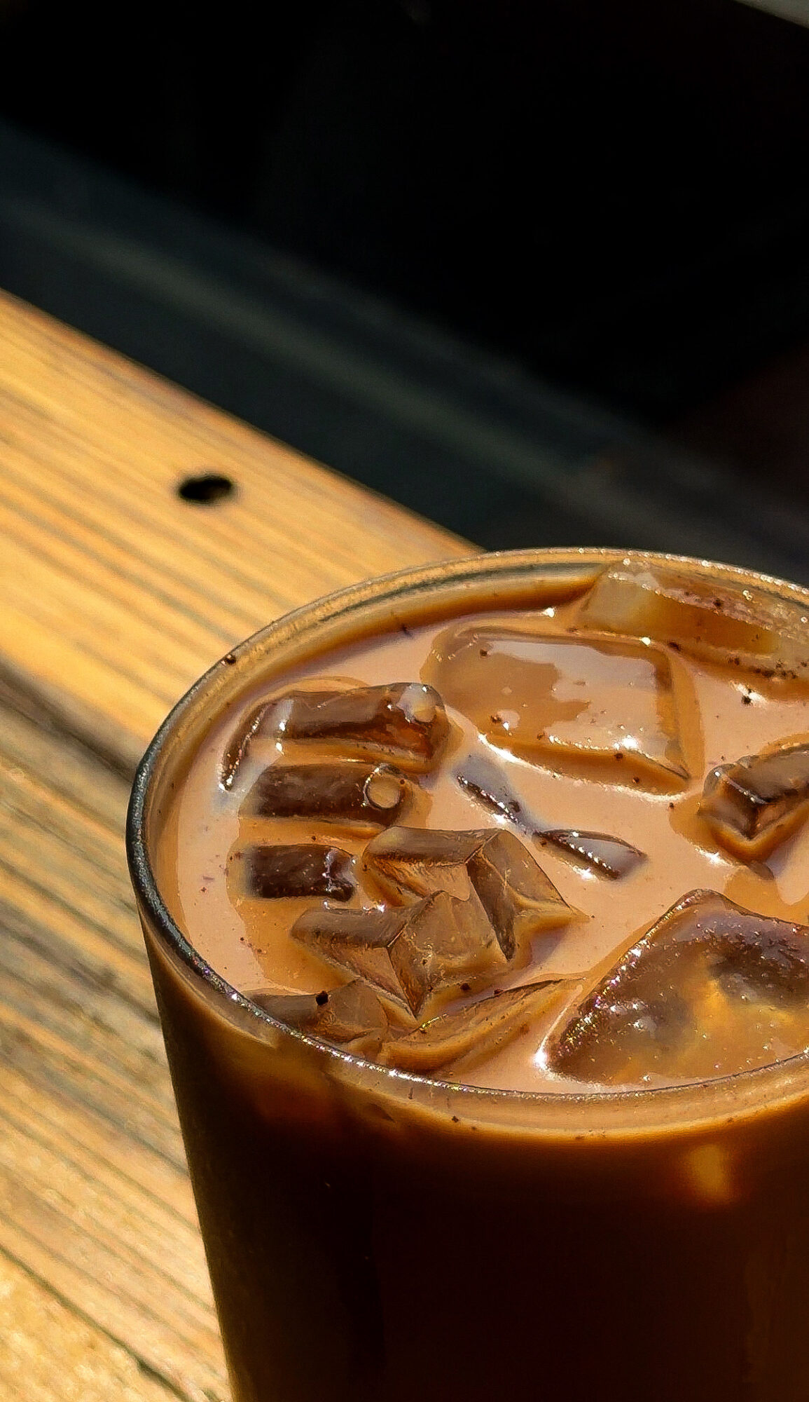 A glass of iced coffee with ice cubes on a wooden surface, viewed from above.