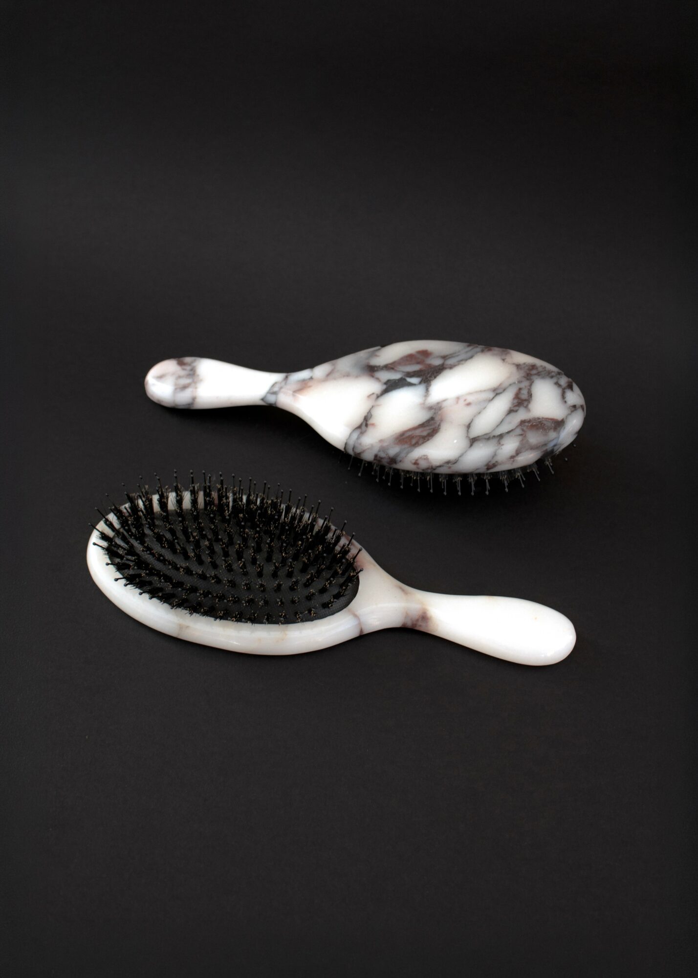 Two hairbrushes with marble-patterned handles and black bristles on a black background.