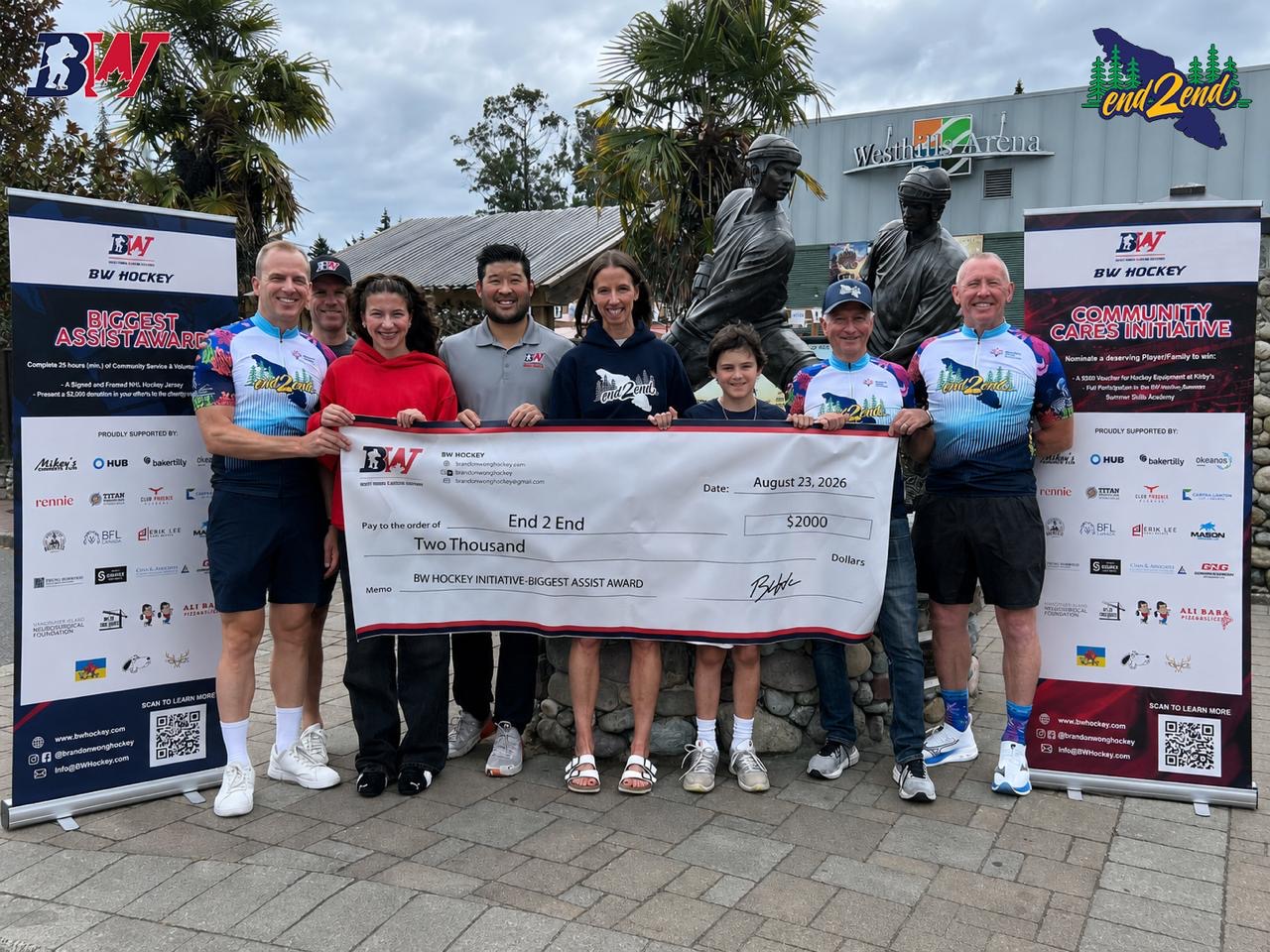 Group of eight people holding a large check in front of a sports venue, with statues and banners in the background.